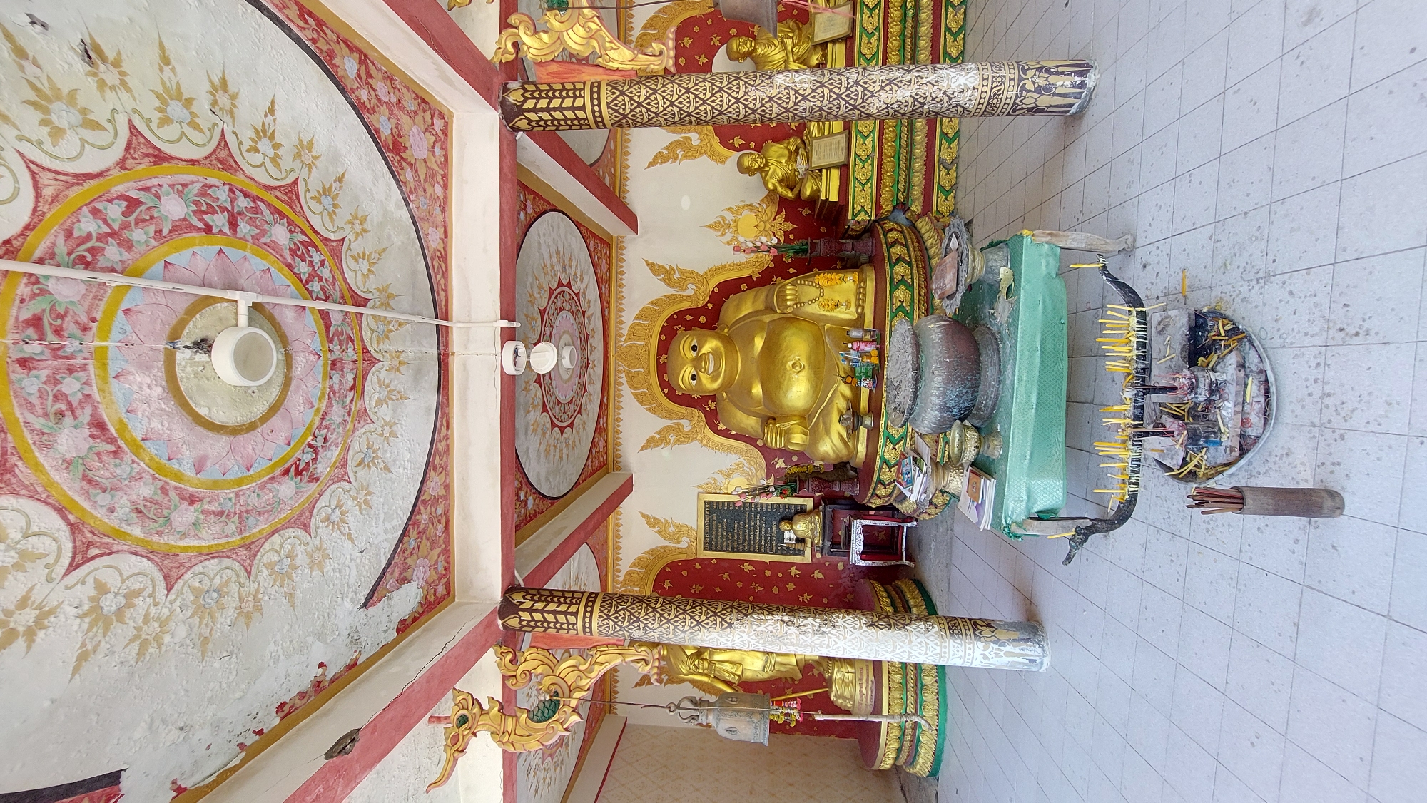 The image displays the vibrant interior of a Buddhist temple or shrine in Baan Plai Laem, Thailand. The central subject is a large, golden, smiling statue of a Buddha figure, likely Hotei or Budai, symbolizing good fortune and abundance. This prominent statue rests on a raised platform, set against a wall richly decorated with intricate golden patterns on a red background.
Surrounding the main figure are several smaller golden Buddha statues and various offerings. In the foreground, a green-draped stand and two large, ornate metallic bowls are visible. On the light-tiled floor to the right, a large circular tray is filled with numerous yellow candles and spent incense sticks, indicating a recent or ongoing act of religious devotion. A wooden holder containing incense sticks stands beside the tray.
Architecturally, the temple features two decorated pillars adorned with gold and white patterns. The ceiling is a notable detail, showcasing large, concentric circular motifs in red, yellow, pink, and white, accented with delicate floral designs. White PVC pipes with light fixtures are installed across the ceiling, providing illumination. Some areas of the painted surfaces show signs of wear and minor cracking. A dark rectangular plaque with unreadable Thai script is mounted below the main Buddha statue. No people are present in the image, contributing to a serene and spiritual atmosphere.