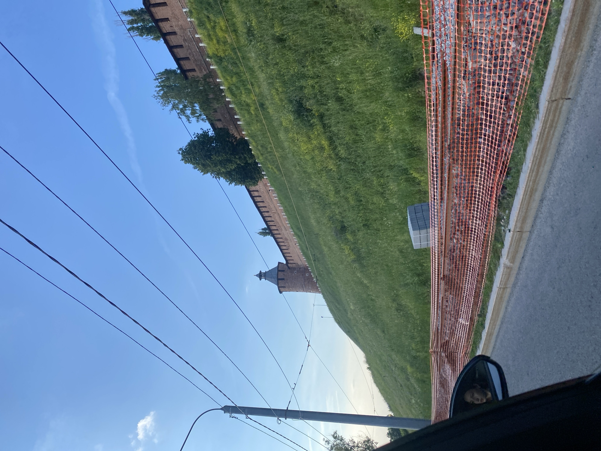 The image depicts a view from a car looking towards a grassy hill with a brick wall and a tower at the top, characteristic of Nizhny Novgorod, Russia. In the foreground, the side mirror of the car reflects a woman's face. An orange construction fence is visible along the side of the road, indicating ongoing work. Overhead power lines stretch across the bright blue sky. The hill is covered in lush green grass, with some yellow wildflowers scattered throughout. The brick wall appears to be a historical fortification, possibly part of the Nizhny Novgorod Kremlin.