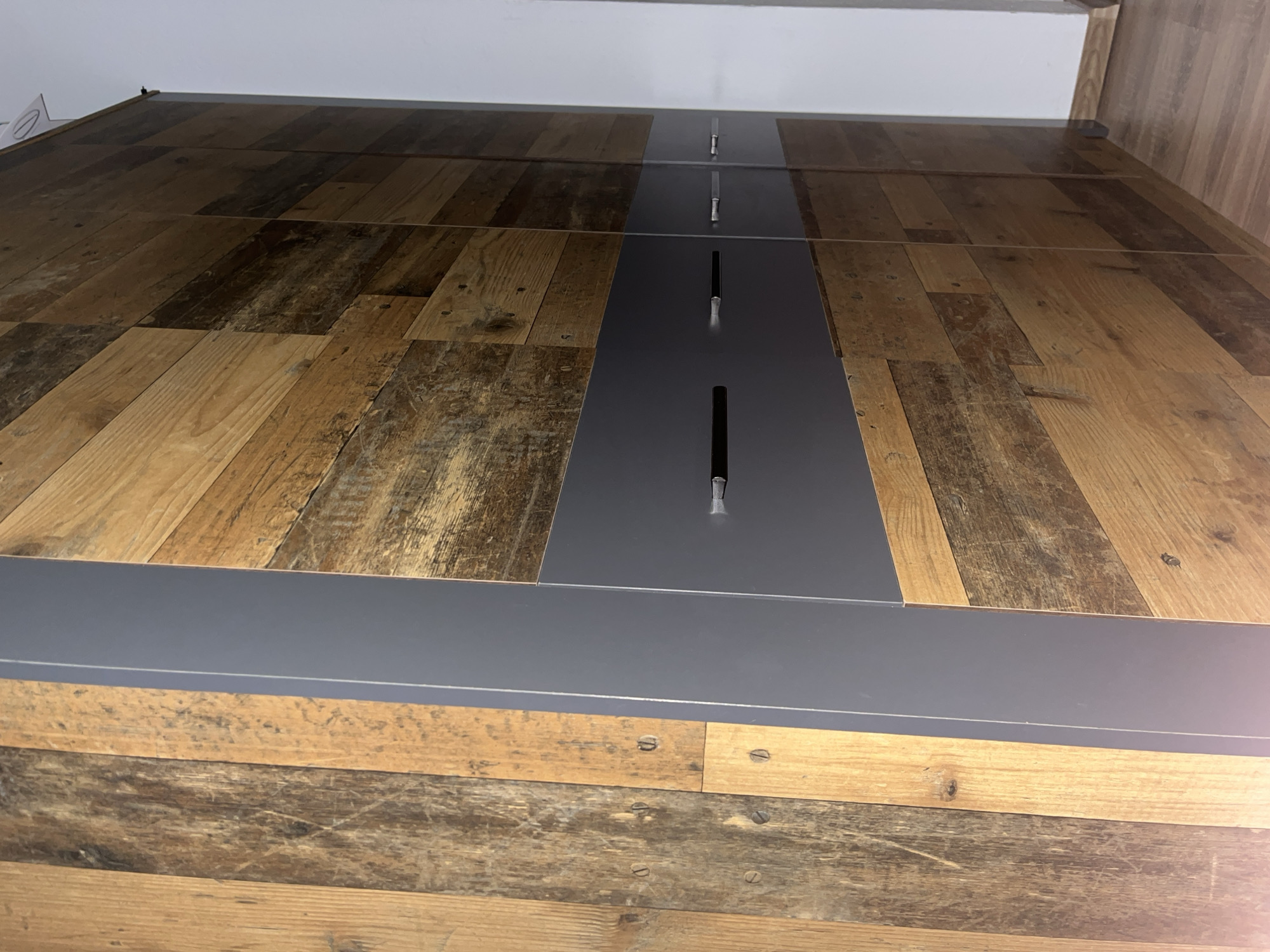 The image shows a close-up, low-angle view of a wooden surface with a gray metal fixture attached to it. The wood appears to be constructed of planks with varied shades, from light brown to darker, almost weathered brown. The gray metal fixture runs lengthwise along the wood surface, with intermittent rectangular openings and metal protrusions. The fixture is attached to the wood with screws, which are visible on the planks. Above the wooden surface, a white ceiling is partially visible. The location is unknown.