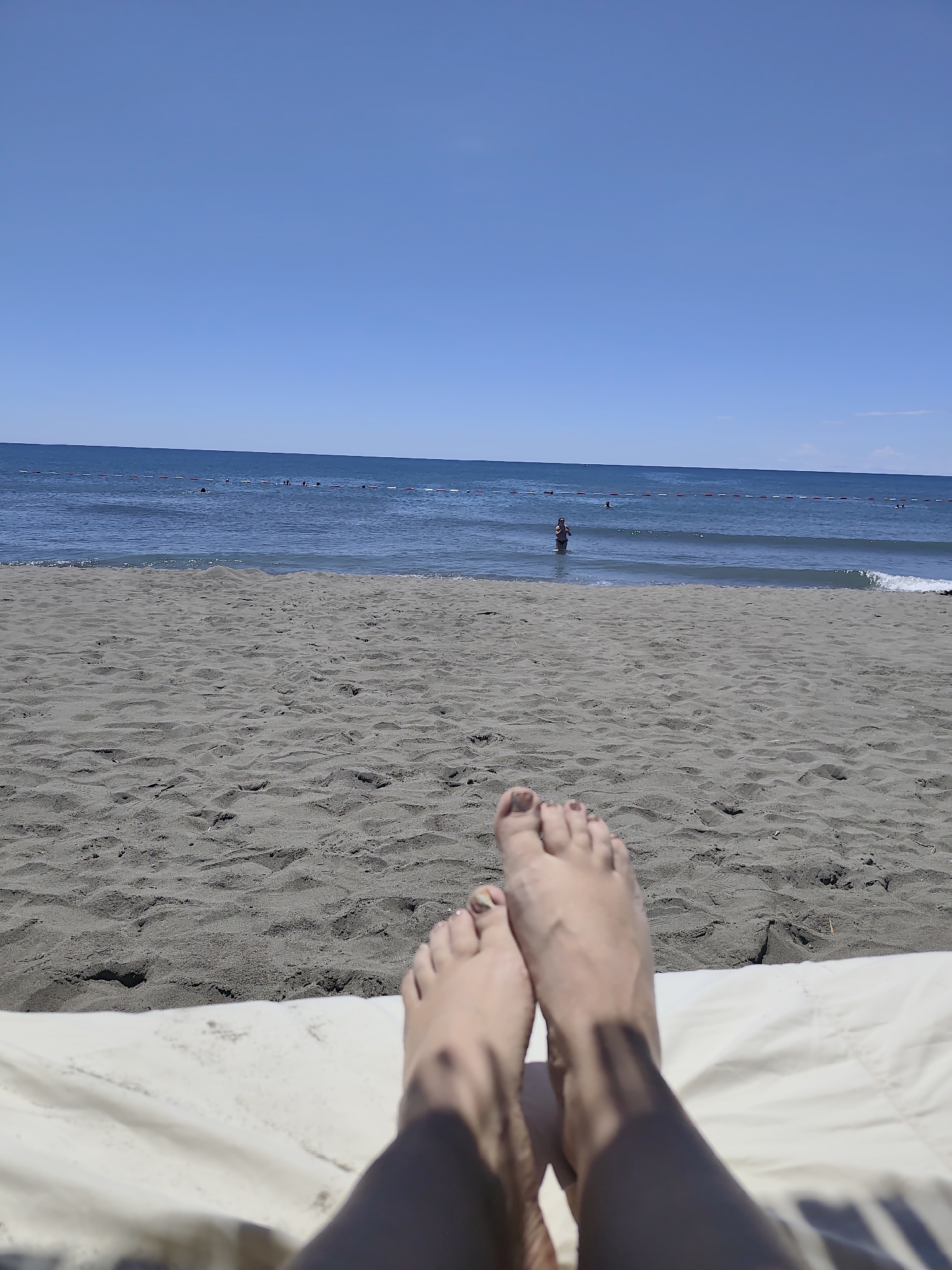This is a first-person perspective photograph taken at a beach in Donji Štoj - Shtoji i Poshtëm, Montenegro. The viewpoint is from someone lying down on a light-colored surface, likely a beach chair or towel, with their feet and lower legs prominently in the foreground. The feet are bare, with painted toenails. The sand in the immediate foreground is a dark grey color and appears somewhat coarse and rippled. Beyond the sand, the blue ocean stretches to the horizon under a clear blue sky. In the water, there are several people visible, some swimming and others standing near a rope barrier that delineates an area of the sea. The lighting suggests it is daytime with bright sunlight.