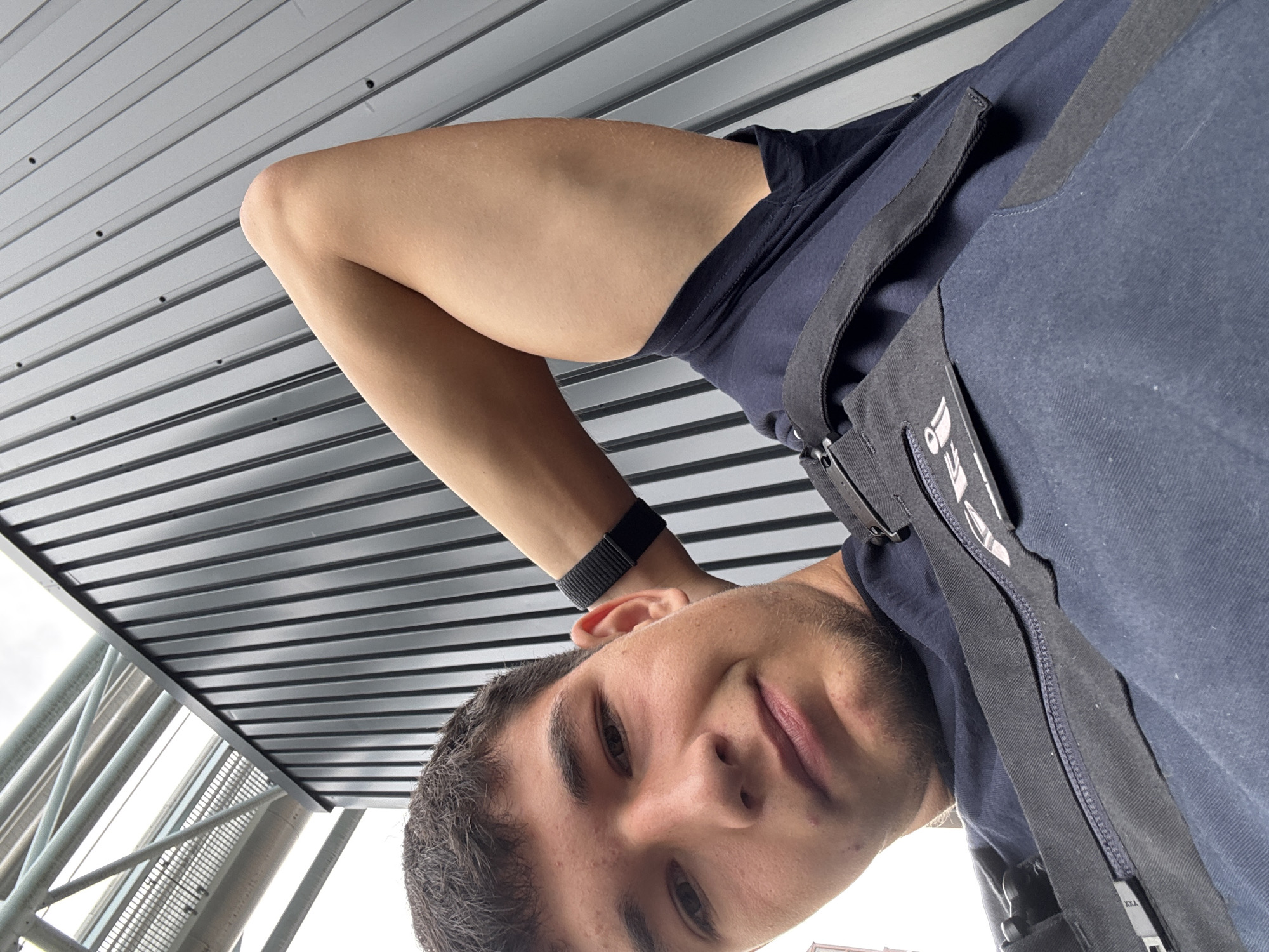 A young man is captured in a close-up, low-angle selfie. He has dark hair and is wearing a dark blue shirt with black straps visible. One arm is raised and resting behind his head, revealing a black band on his wrist. The background consists of a modern, dark gray corrugated metal wall and some metallic infrastructure overhead. The overall setting appears to be an urban industrial or construction area.