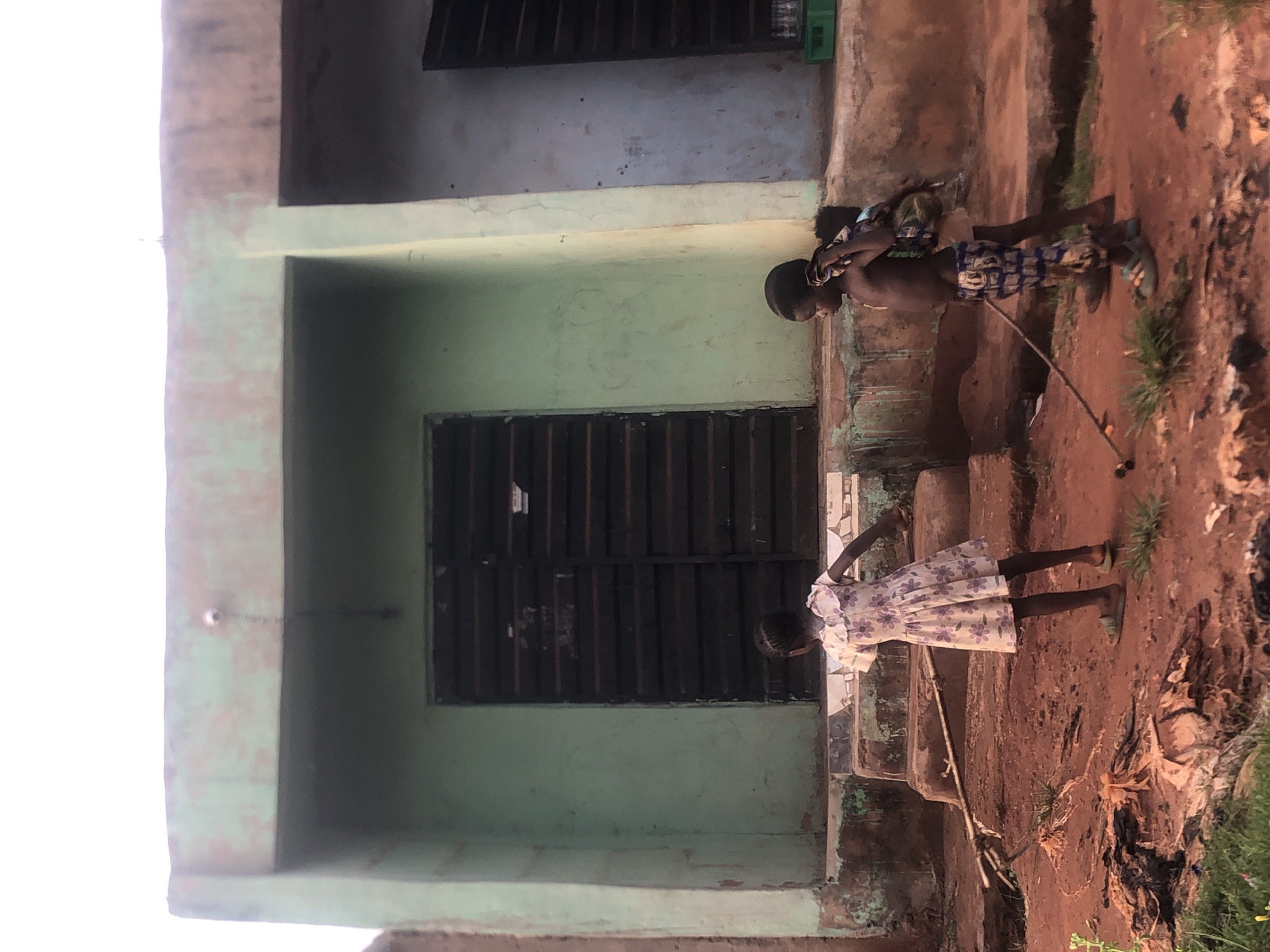 This image, set in Ukwu-Abwa, Nigeria, captures a scene outdoors with two children near a building.
**Subjects:** The primary subjects are two young children, one boy and one girl. The boy, shirtless and wearing patterned shorts, appears to have a younger child or a bundle strapped to his back. He is holding a long stick. The girl is wearing a light-colored dress with a floral pattern and is standing on steps. The main building in the background has light green/teal walls and dark, louvered windows or shutters. The ground is reddish-brown earth, with some sparse green vegetation and scattered debris.
**Activity or event taking place:** The children appear to be engaged in everyday activities. The boy is walking or standing with his stick, possibly playing or using it for support. The girl is standing on the steps, looking down, suggesting she might be observing something or waiting.
**Notable details:** The scene depicts a rural or semi-urban environment. The building has a weathered appearance. The ground is characteristic of laterite soil common in tropical regions. The lighting suggests it is daytime, though the exact time is not precisely discernible due to soft shadows and bright, diffused light. There is no visible text in the image. The children's expressions are not clearly visible, but they seem casually engaged in their surroundings.