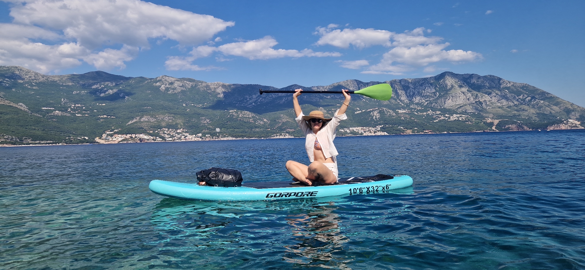 A person is paddleboarding on a bright blue, clear body of water with rolling green mountains in the background. The person is sitting cross-legged on the paddleboard, holding a paddle above their head with both hands. They are wearing a straw hat, sunglasses, a white button-up shirt, and white shorts over a striped bikini. The paddleboard is turquoise and black, with "GORPORE" written on the side. A black dry bag is secured to the front of the paddleboard. The water is calm with subtle ripples, and sunlight reflects off the surface. The sky is a vibrant blue with scattered white clouds. The mountains behind are covered in lush green vegetation and some rocky outcrops, with scattered buildings visible along the coastline. The scene appears to be in a coastal area near Boreti, Montenegro, given the characteristic landscape.