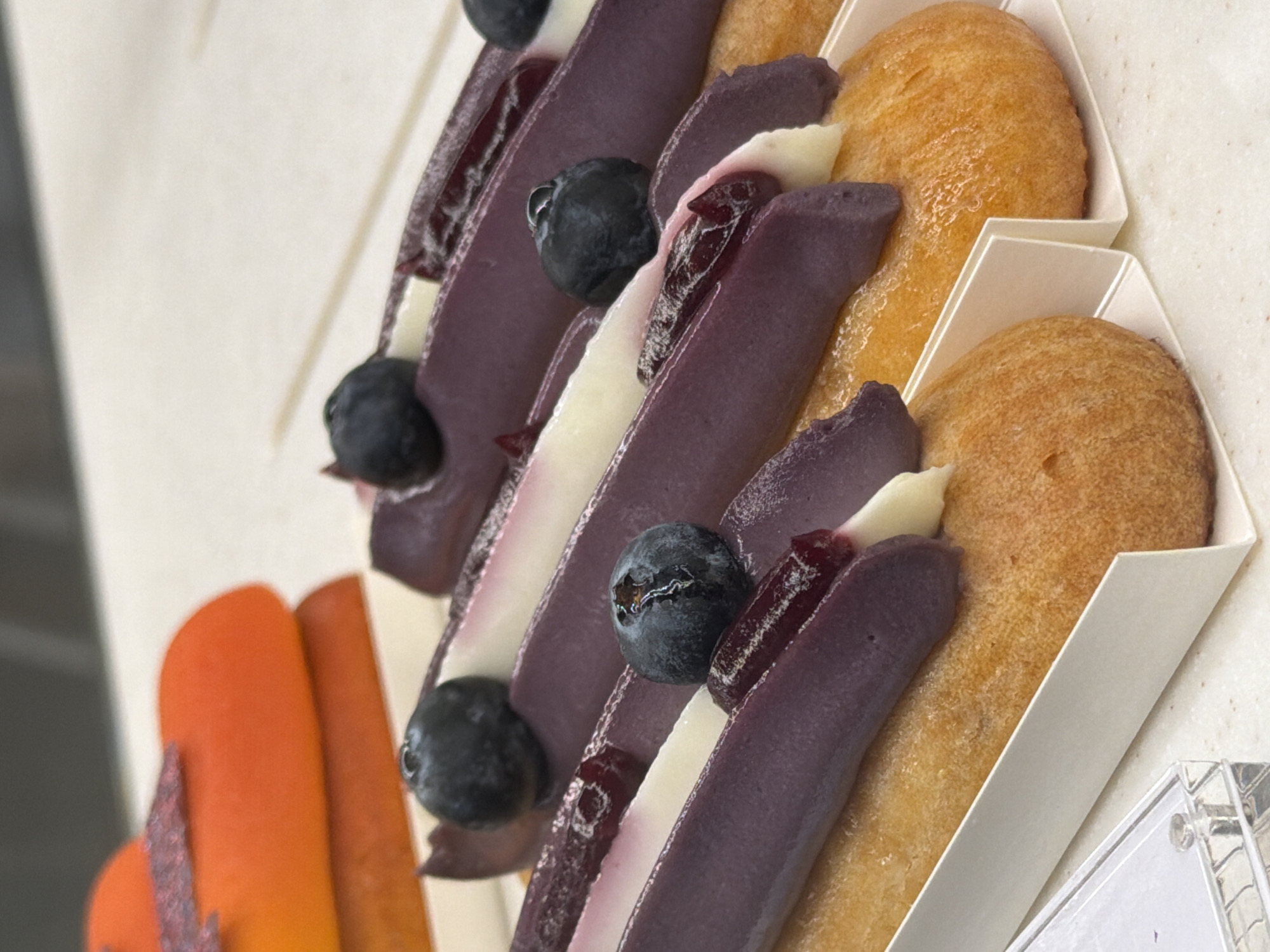The image showcases a close-up, angled view of several small pastries arranged in white paper holders. The pastries appear to be individual servings, lined up diagonally.
The subjects are primarily food items. There are several purple-colored pastries, possibly éclairs or cream puffs, topped with a white cream, blueberries, and a dark jam or filling. Some of these purple pastries also have a glistening red element. Alongside these, there are golden-brown, puffed pastries that resemble small buns or brioches. In the foreground on the left, there are two orange-colored elements, one with a textured, possibly glittery, surface.
The setting seems to be a display or serving area for desserts, likely in a bakery or cafe. The lighting is bright and even, suggesting an indoor environment. There are no visible people, and the focus is solely on the presentation of the food. The composition is tight, with the pastries filling most of the frame, giving the viewer a detailed look at their textures and toppings. The white paper holders provide a clean, minimalist contrast to the vibrant colors of the food. No text is visible in the image. The overall impression is one of finely crafted, visually appealing desserts.