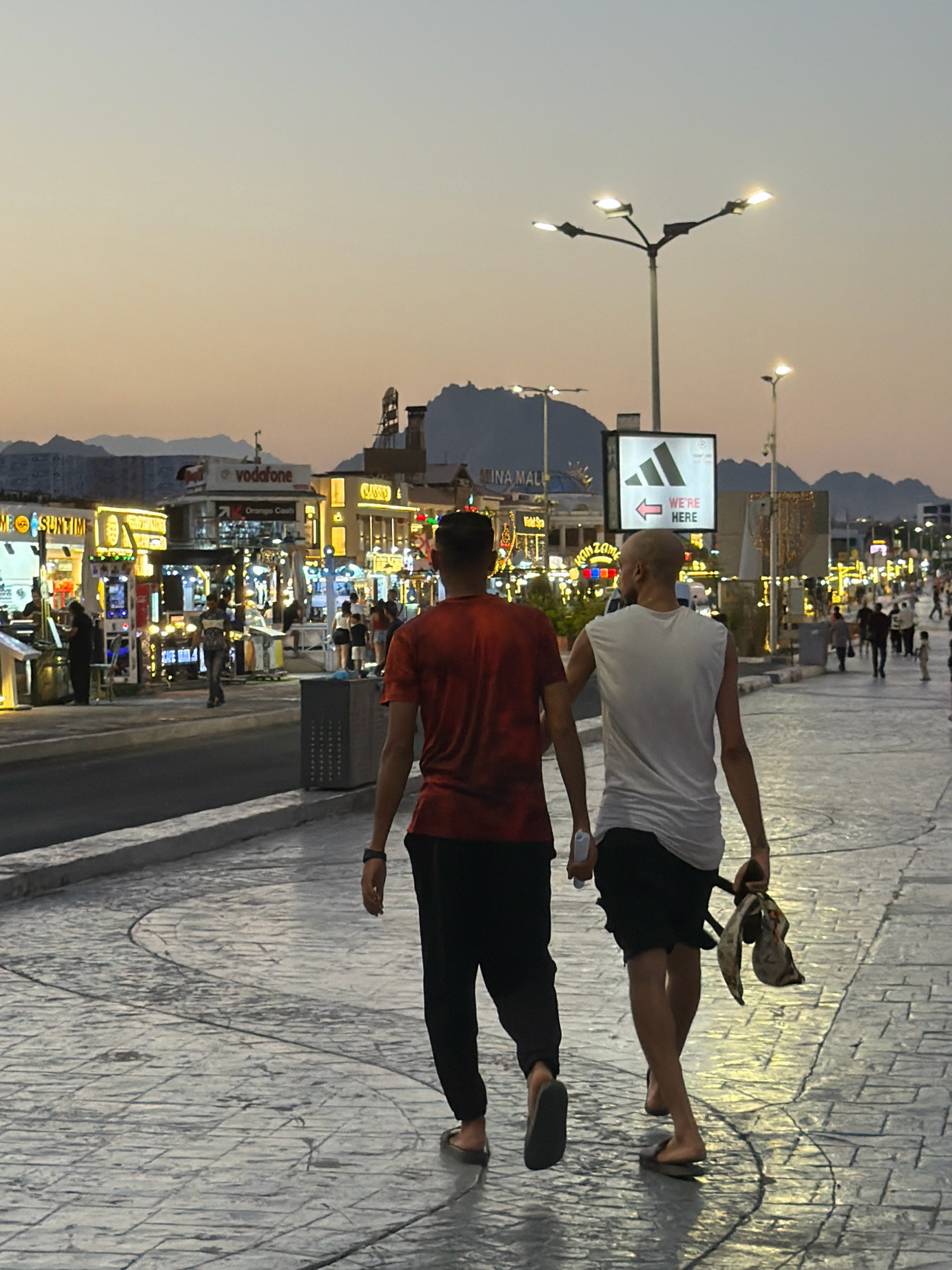 Two individuals are walking away from the camera on a pedestrian walkway lined with shops and streetlights. The person on the left is wearing a red t-shirt and black pants. The person on the right is wearing a white sleeveless shirt and black shorts, carrying a patterned cloth bag and a piece of paper. They are walking on a patterned stone walkway. In the background, there are buildings with illuminated signs, including "vodafone," "Orange Cash," and "CLASSICO." Mountains are visible in the distance under a twilight sky. The scene appears to be in Sharm El Sheikh, Egypt, given the presence of many tourist-oriented establishments and the distinctive mountainous landscape. The overall atmosphere is that of a busy evening stroll in a commercial area.