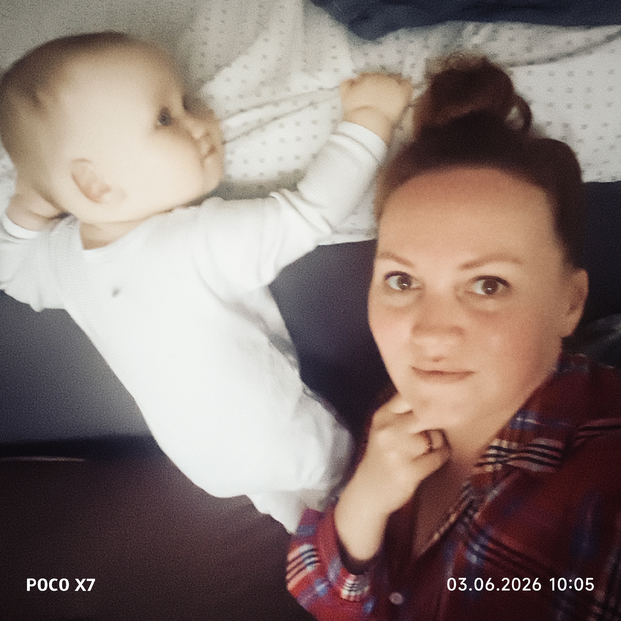 This media file features a candid, slightly blurry selfie showing a woman and an infant. The scene appears to be indoors, likely a bedroom, with both subjects resting on white bedding with a subtle dotted pattern.
The woman, visible in the foreground on the right, is looking directly at the camera. She has light-colored hair pulled back, visible brows and eyes, and a neutral to slightly content expression. She is wearing a red and blue plaid shirt, and her hand, wearing a ring on one finger, is gently resting near her chin.
To the left and slightly behind the woman is a baby, appearing to be an infant or young toddler, lying on its back. The baby is dressed in a white, long-sleeved bodysuit or sleepsuit and is looking off to the left side of the frame, away from the camera. Its face and body are slightly out of focus compared to the woman's.
The image has a soft, grainy quality, suggesting it might have been taken in low light or with a filter applied. The overall tone is intimate and casual, capturing a moment between a parent and child.
Visible text in the bottom left corner reads "POCO X7", indicating the device used to take the photo. In the bottom right corner, the date and time "03.06.2026 10:05" are displayed.