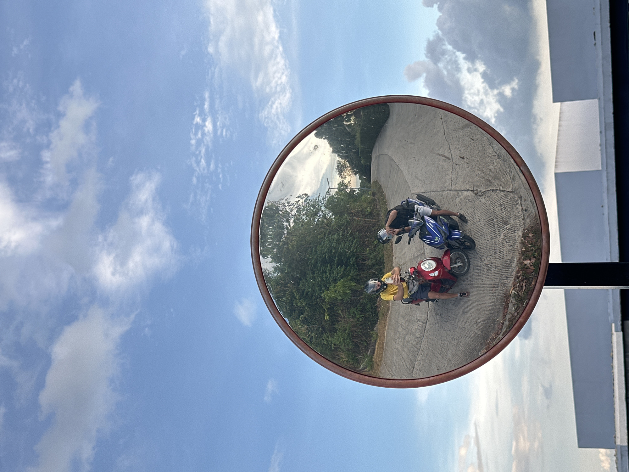 An eye-level outdoor photograph shows a circular convex mirror reflecting two men on motorbikes parked on a paved road next to a wooded area. The men are facing each other with their motorbikes between them. The first man, wearing a yellow shirt and a helmet, is holding a phone in his hand. The second man, wearing a black shirt, light-colored shorts, and a helmet, is standing near his blue and black motorbike. The mirror is attached to a black pole affixed to a white structure with gray accents. In the background, the sky is blue with wispy clouds. The setting is Baan Thong Po, Thailand.