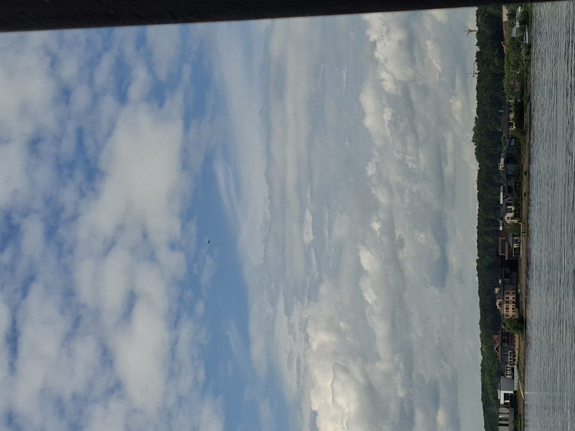 A body of water is in the foreground, with a shoreline visible in the middle ground. On the shoreline, there is a line of houses and trees. In the background, the sky is filled with clouds. A small dark object, possibly a bird or drone, is visible in the upper portion of the sky. The image appears to have been taken from a boat or from the water's edge, looking towards the shore. The architectural style of the houses suggests a modern residential area. The presence of trees indicates a natural or semi-natural environment bordering the development. The sky with clouds implies it is daytime. The image is of moderate quality, with some graininess.