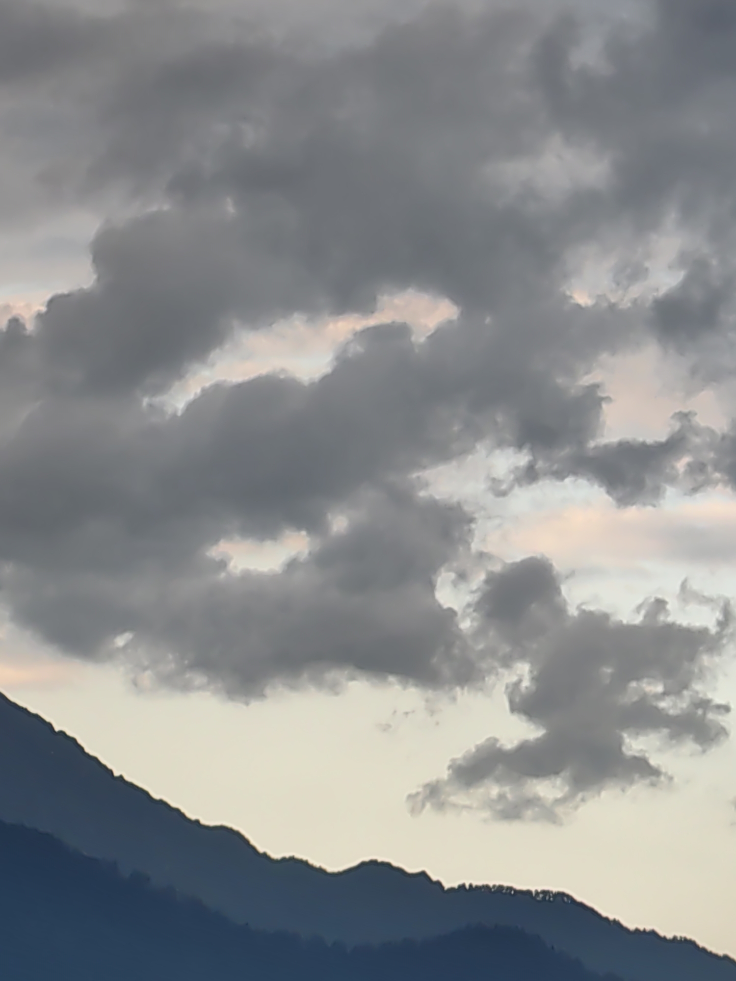 The image shows a mountain range at dusk. The sky is filled with dark, dramatic clouds, with patches of soft pink and orange light peeking through. The mountains are silhouetted against the sky, their peaks appearing sharp and jagged. The lower slopes of the mountains are a deep blue, while the upper slopes are a lighter shade of gray. The overall mood of the image is serene and peaceful, with a sense of the vastness and power of nature. The scene is likely Vashisht, India, a popular tourist destination known for its natural beauty and hot springs.