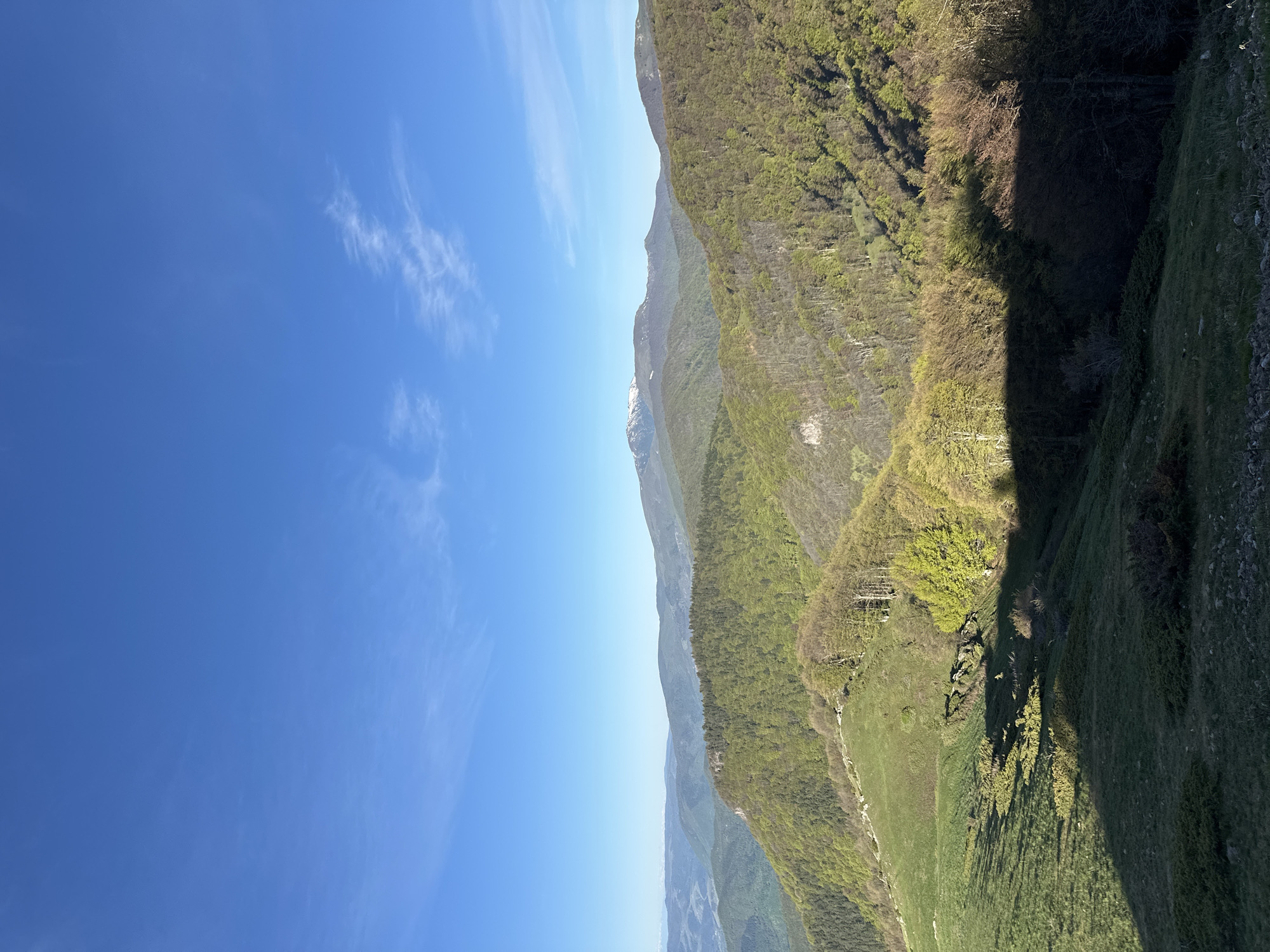 The image presents a landscape view, potentially of mountainous terrain, under a clear blue sky with scattered wispy clouds. The right side of the image is dominated by a forested hillside or mountain slope covered with various shades of green vegetation. Shadowing obscures some of the lower portions of the hillside. In the background, there are other mountain ridges. The area appears to be a remote natural location, and the context suggests it might be near Hristo Danovo, Bulgaria.