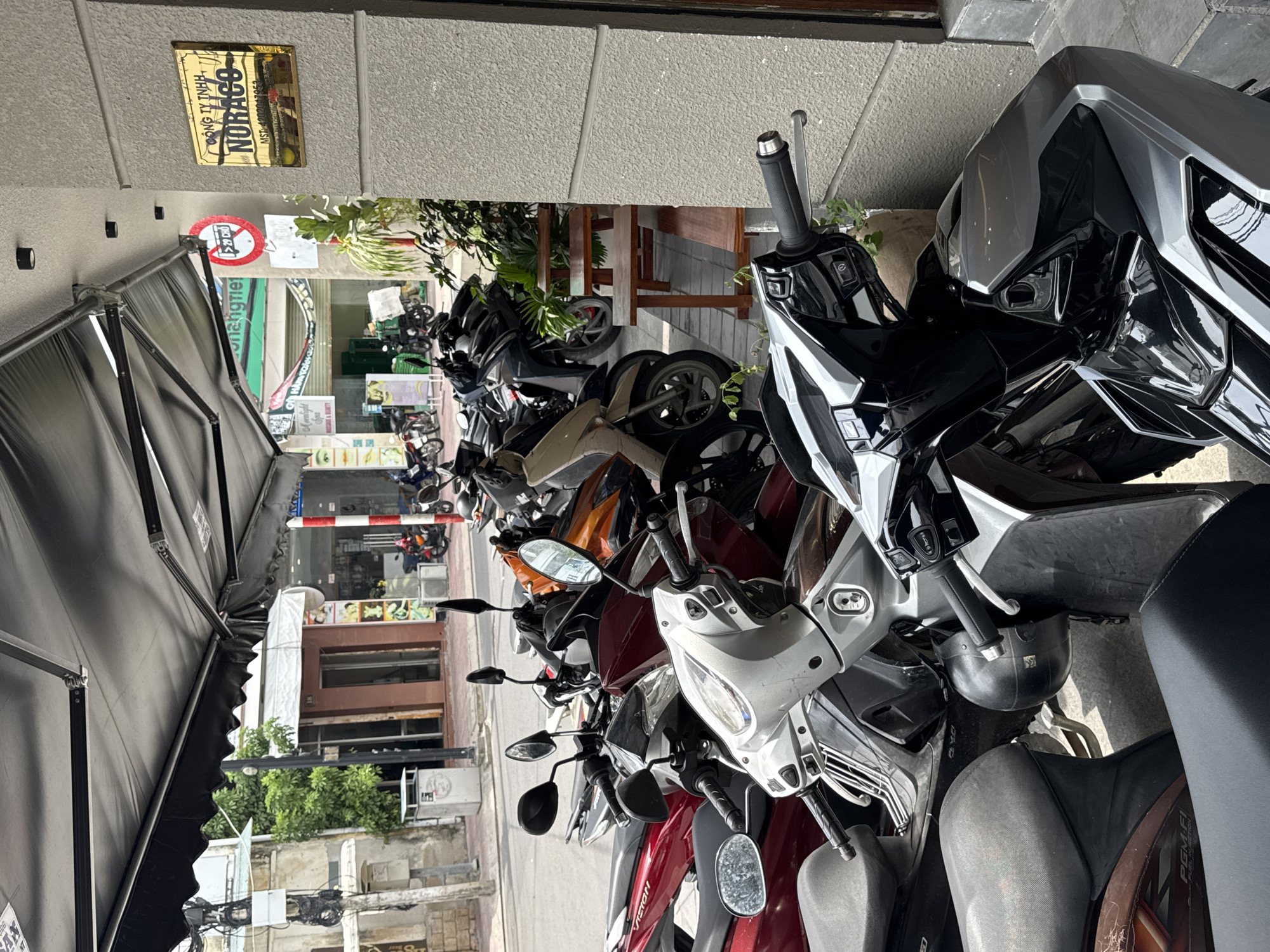 The image shows a row of parked motorcycles outside a building with a gold plaque that reads "CONG TY TNHH NORAGO MST: 4200047053". A black awning extends over the area where the motorcycles are parked. In the background, there are buildings, trees, and other motorcycles. The setting appears to be a street in Nha Trang Ward, Vietnam. There are no people visible in the image. The motorcycles are various colors and models, and they are all parked closely together. The building in the background is a light gray color, and it has a glass door. The street is paved with concrete. The overall scene depicts a common sight of motorcycle parking in Vietnam.