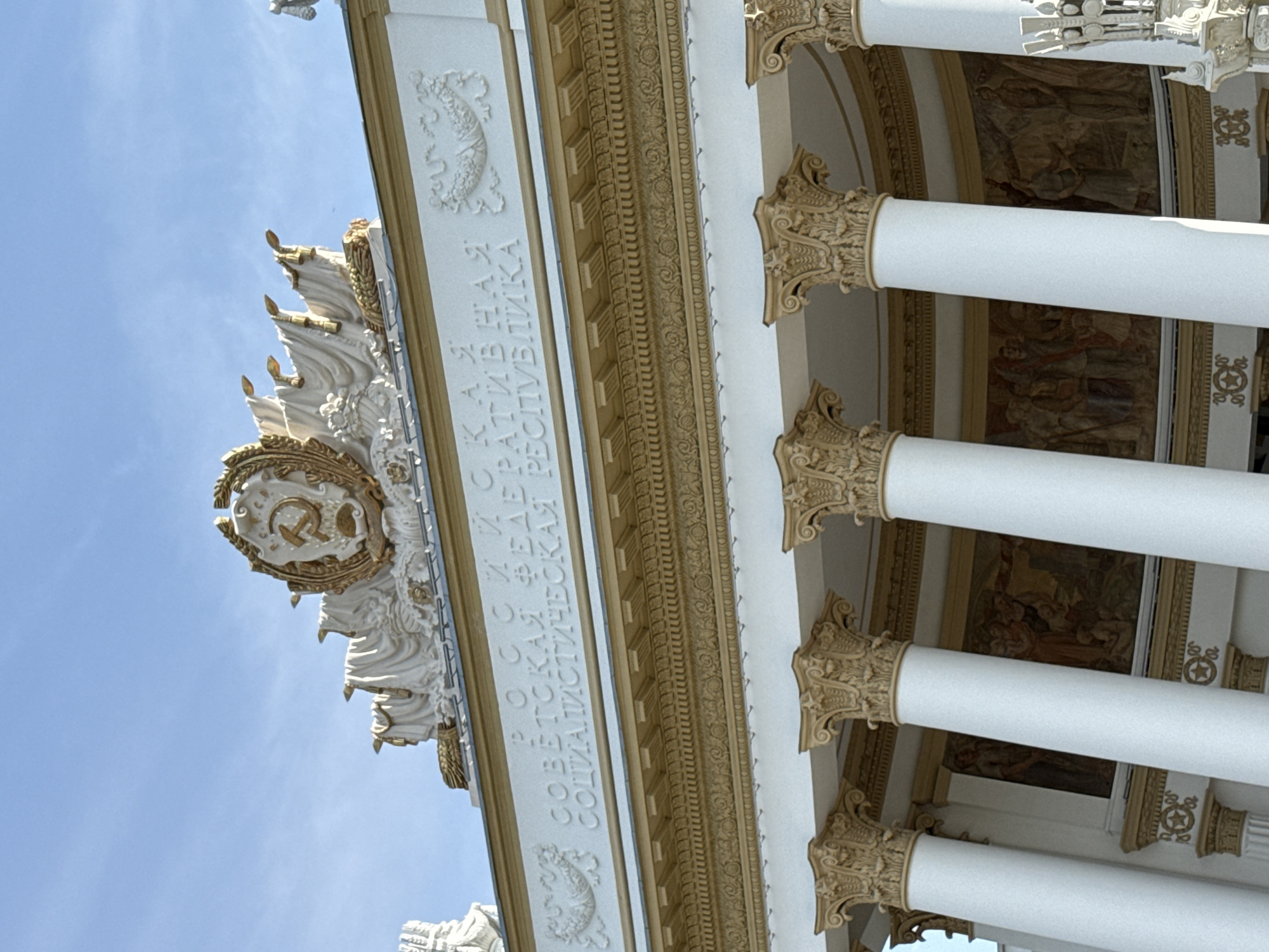 The image shows a portion of a building in Moscow, Russia. The building features white classical columns with ornate, gold-colored capitals and a decorative entablature. The Soviet hammer and sickle symbol is prominently displayed in a golden medallion. Inscribed vertically next to the medallion is Russian text. Murals can be seen in the spaces between the columns and above the entablature. The background is a light blue sky.
