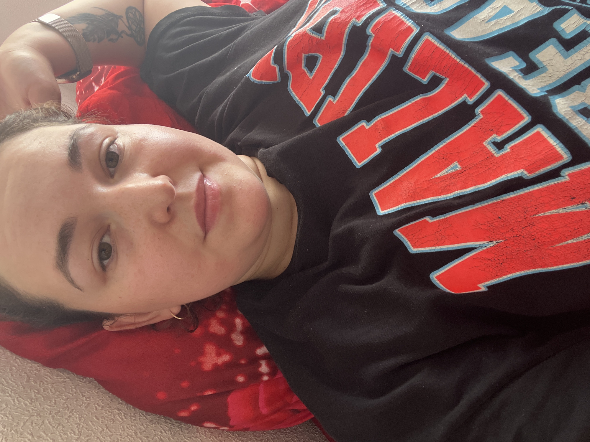 The image shows a close-up, selfie-style shot of a person lying down with their head resting on a red, patterned pillow. The person is wearing a black sweatshirt with large, distressed red and blue lettering on the front. The person's face is partially visible, and they appear to have light skin with some freckles. Their right arm is raised behind their head, and a tattoo is visible on their upper arm. The background is out of focus and appears to be a textured wall. There are no other individuals or objects in the frame that provide any context for the location being in the city of Chelyabinsk, Russia.