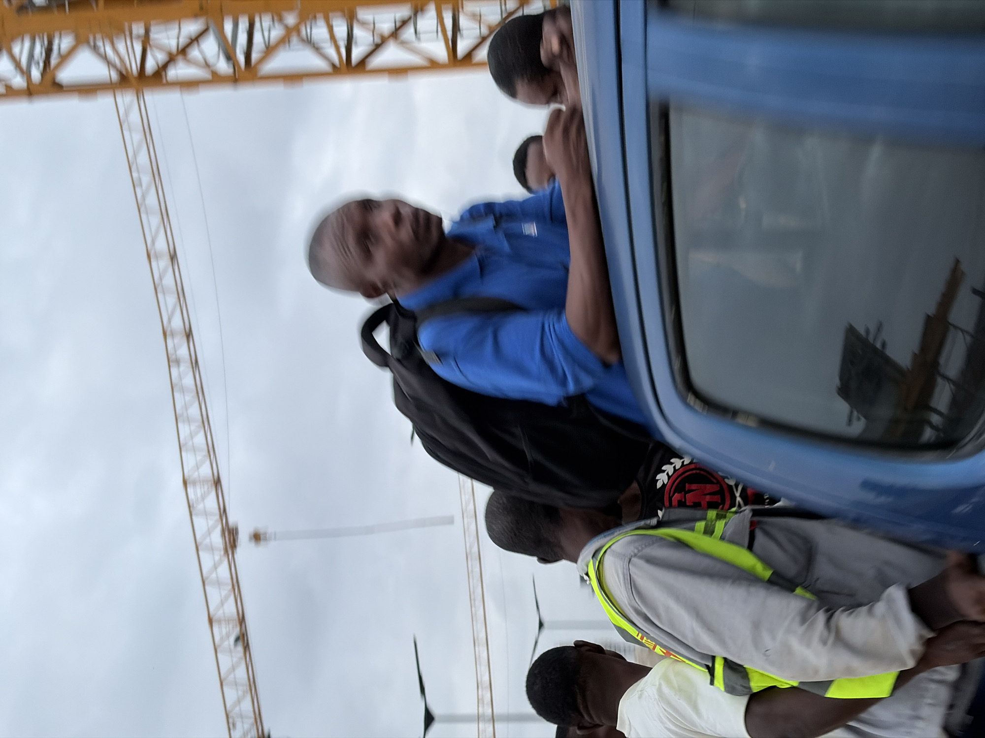 The image depicts a group of men in an outdoor urban environment, likely in Alaguntan, Nigeria, as indicated by the presence of construction cranes. The sky is overcast. In the foreground, a man wearing a blue polo shirt and a backpack is leaning over the roof of a blue vehicle. Beside him, another man is wearing a gray work jacket with a high-visibility yellow vest. To the left, a man in a white t-shirt is partially visible. Other individuals are also present, some appearing to be inside the vehicle or near it. The overall scene suggests a moment of movement or gathering, possibly related to transportation or a work site, given the construction equipment in the background.