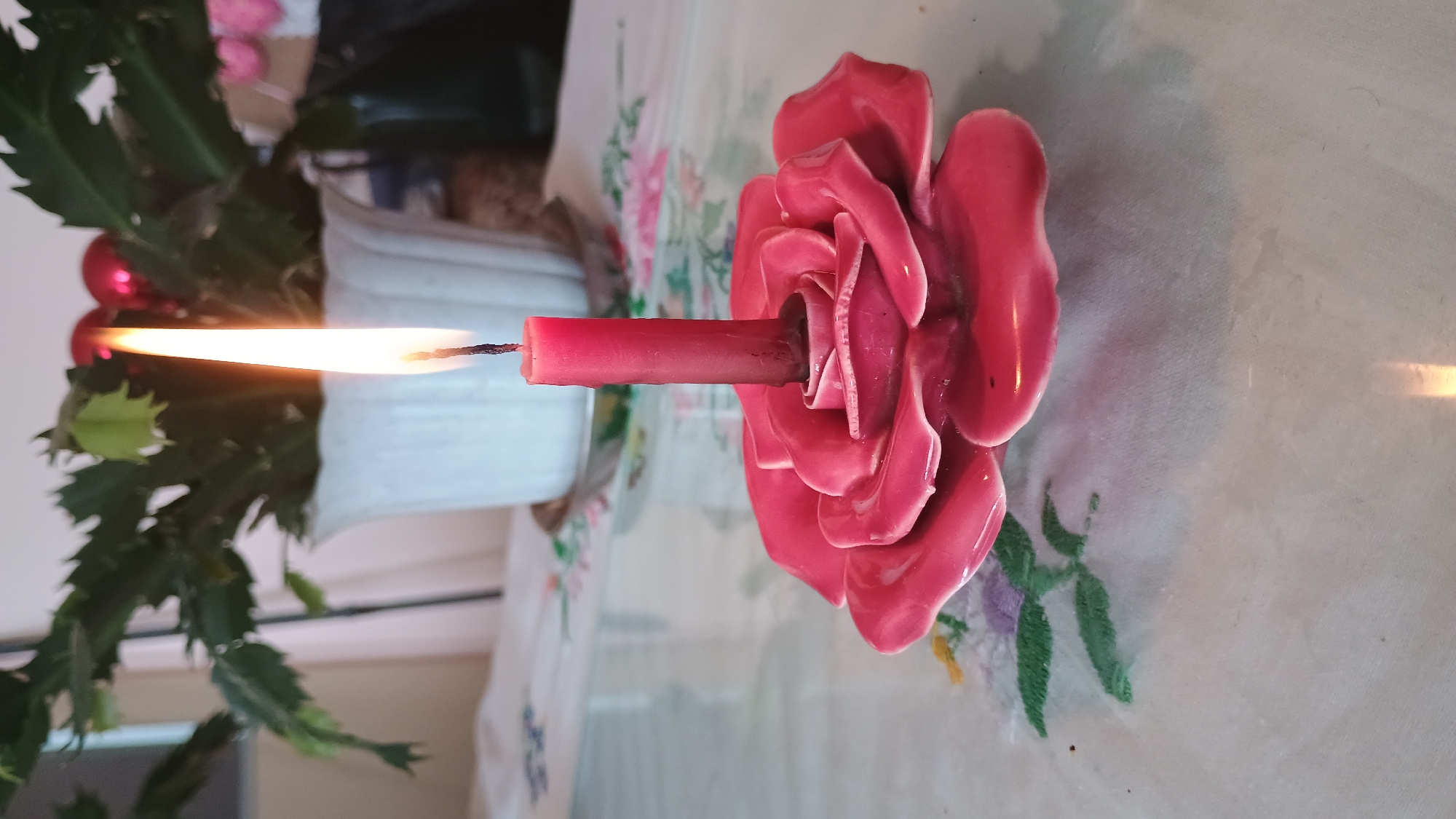 A lit red candle in a rose-shaped ceramic holder rests on a reflective surface, likely a table. The candle is tall and slender, with a visible flame. The rose holder is a vibrant pink with detailed petal formations. In the background, a potted plant with green, segmented leaves sits behind the candle, and a white ceramic pot is visible. The setting appears to be indoors, with a tablecloth visible under the reflective surface, which has embroidered floral patterns. The scene is illuminated by the candle's flame and ambient light. The location is not discernible from the image itself.