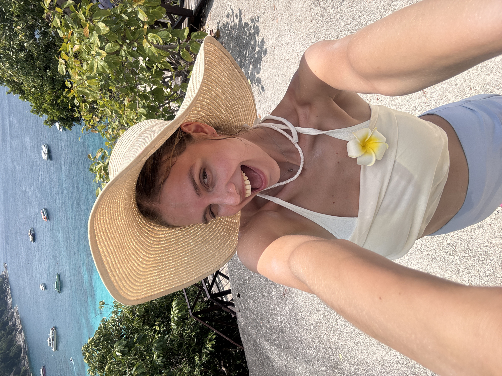A young Caucasian woman is taking a selfie outdoors. She is wearing a large straw hat, a light-colored top with a white flower pinned to it, and light blue bottoms. She is smiling broadly with her mouth open. The background features a blue body of water with boats, lush green vegetation, and a gray concrete surface. The location is the city of Lam Kaen, Thailand.
