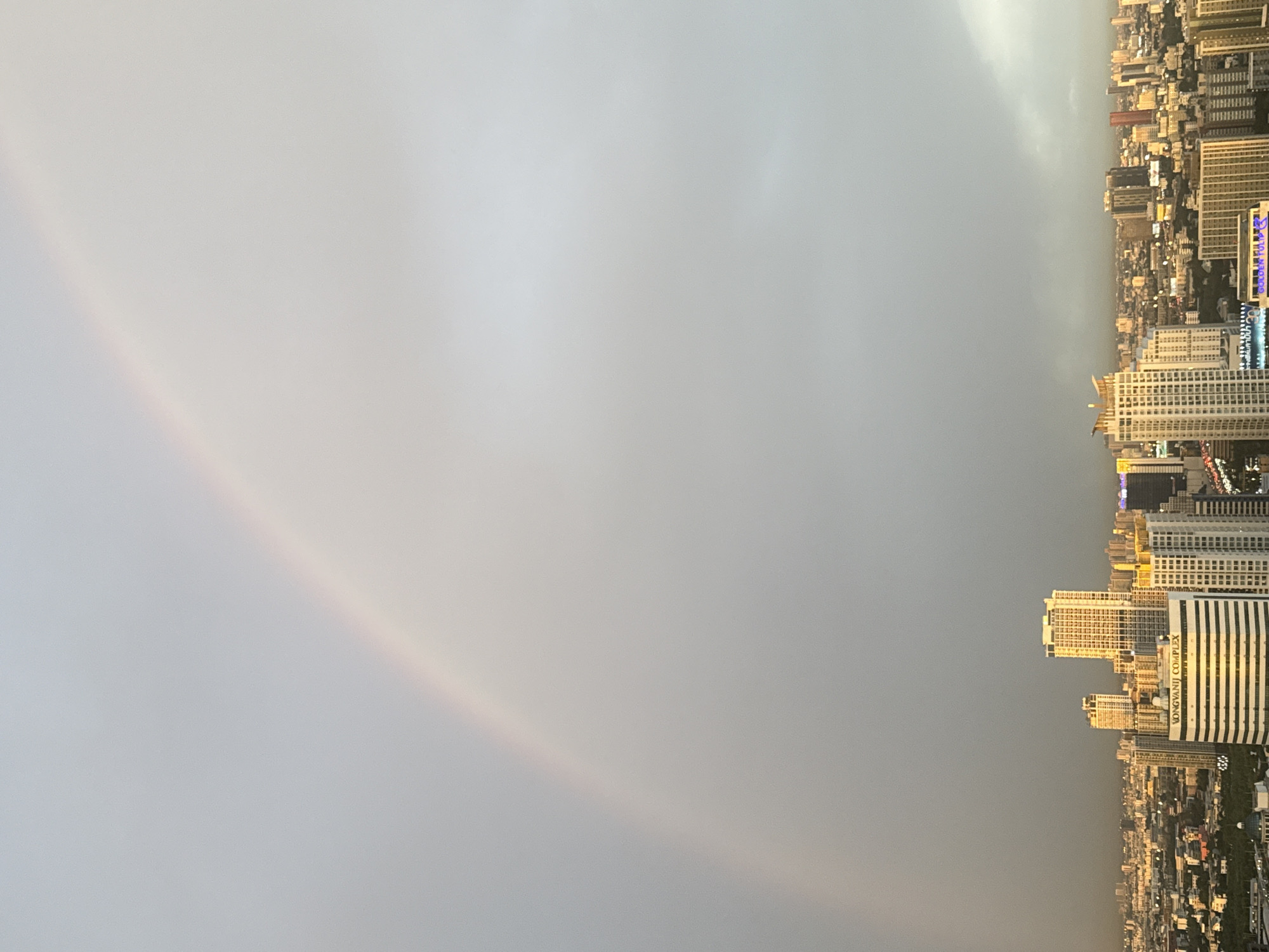 This is a tilted, vertical shot of a cityscape with a rainbow. The scene is dominated by a vast expanse of grey sky, with a faint rainbow arc visible in the upper left quadrant. Below the sky, a dense urban landscape stretches out, filled with numerous tall buildings of varying architectural styles and heights. The buildings are predominantly modern, with many appearing to be residential or commercial complexes.
Some buildings are brightly lit, suggesting it is either late evening or early morning, or that interior lights are on. The cityscape appears to be a sprawling metropolis with a high density of structures. The time of day is unclear due to the overcast sky and the tilt of the image, but the lighting on the buildings hints at a time when artificial lights are beginning to be noticeable. The weather appears to be transitioning, with the presence of a rainbow suggesting recent rain or atmospheric conditions conducive to its formation, followed by clearing skies.
Notable details include the distinct arc of the rainbow, its colors subtly visible against the grey sky. The sheer number and height of the buildings point to a major city. In the foreground, a white building with dark horizontal stripes is clearly visible. Text is visible on one of the buildings in white lettering against a dark background: "VONGVANJI COMPLEX". Given the provided location context of Bangkok, Thailand, this is likely a view of the city from a high vantage point, perhaps from a skyscraper or a viewpoint overlooking the urban sprawl. The image evokes a sense of scale and the coexistence of natural phenomena like rainbows with the built environment of a modern city.