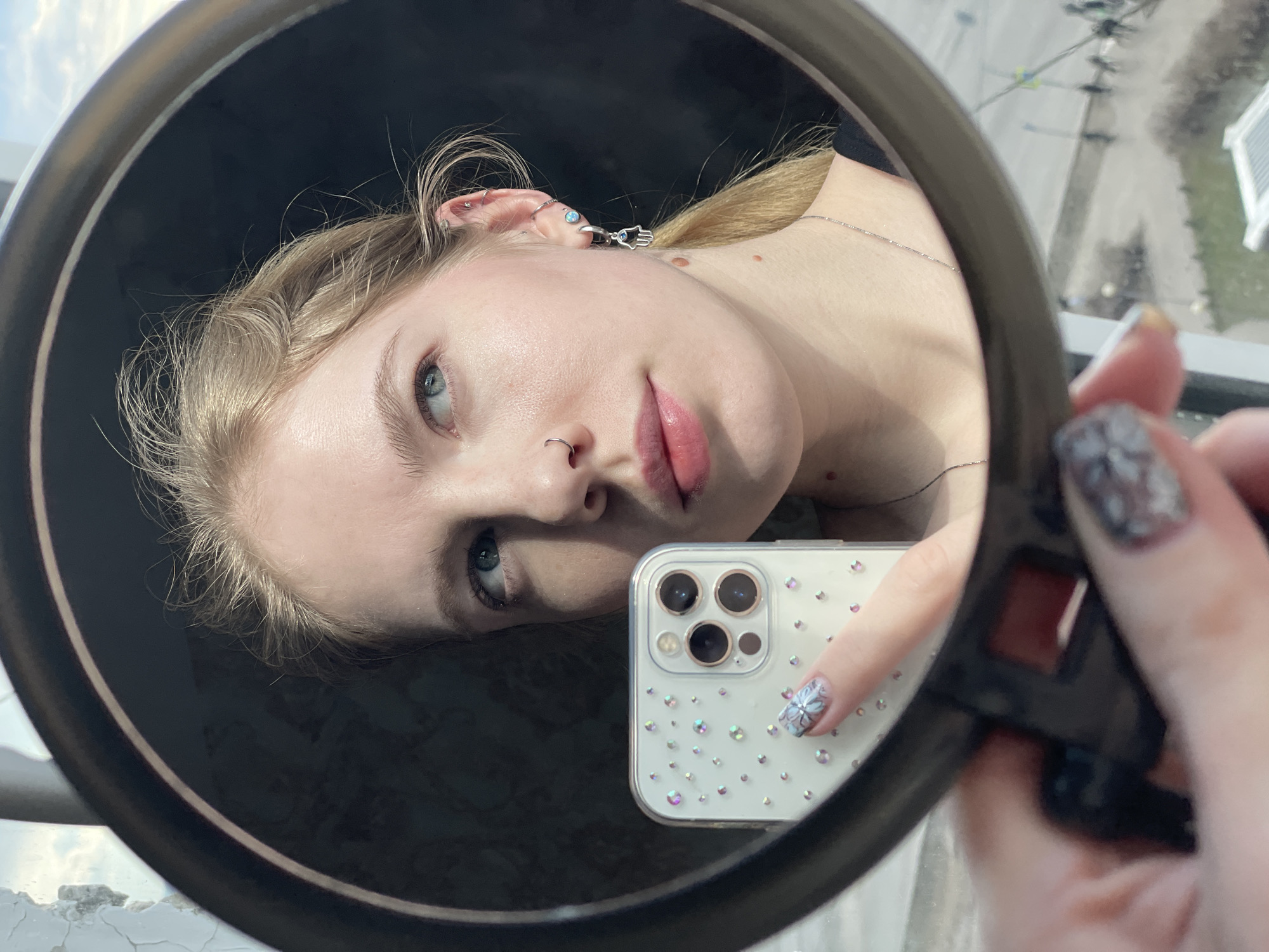 A mirror selfie captures a young woman with light blonde hair, fair skin, and light blue-grey eyes, looking calmly into a round mirror. She has a nose ring and multiple ear piercings, including one featuring a Hamsa hand charm. Her lips are lightly colored with pink lipstick.
Reflected in the mirror is a white iPhone, held by her hand, encased in a clear cover adorned with numerous small, sparkling rhinestones and showcasing a triple camera system. The hand holding the mirror features nails painted with a glittery, patterned design, notably a floral or snowflake motif on the index finger.
The mirror has a dark, circular frame. The background visible outside the mirror's frame suggests an urban or suburban landscape viewed from a high elevation, with buildings and open sky under bright daylight conditions. No visible text is present.