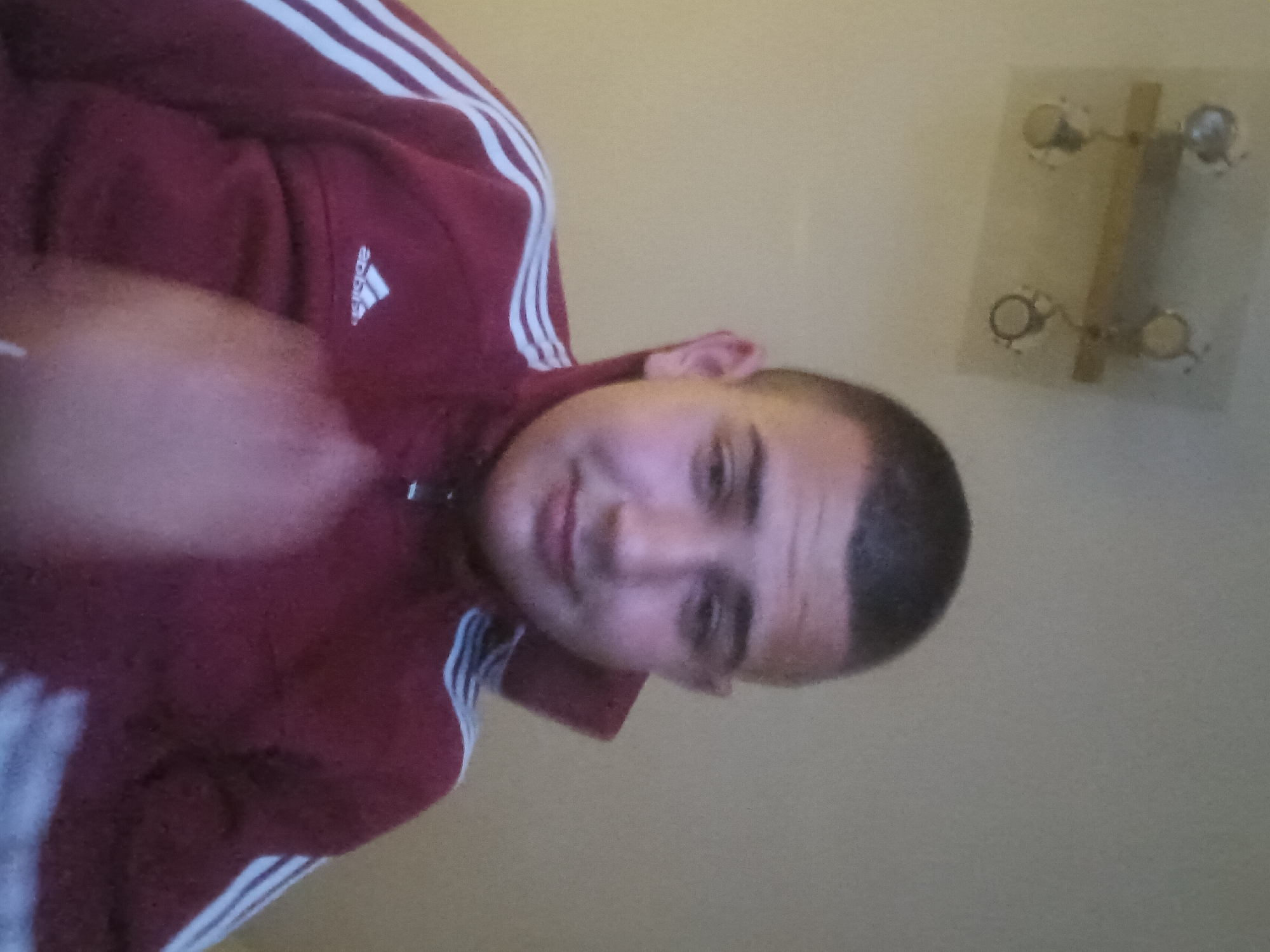 The image shows a man with a buzz cut wearing a maroon Adidas track jacket with white stripes on the sleeves. The background is a plain wall with a small decorative object that appears to be a light switch plate cover with a wooden bar and metallic circular elements. The man is looking towards the camera and appears to be smiling slightly. The city location is Unknown, Unknown.