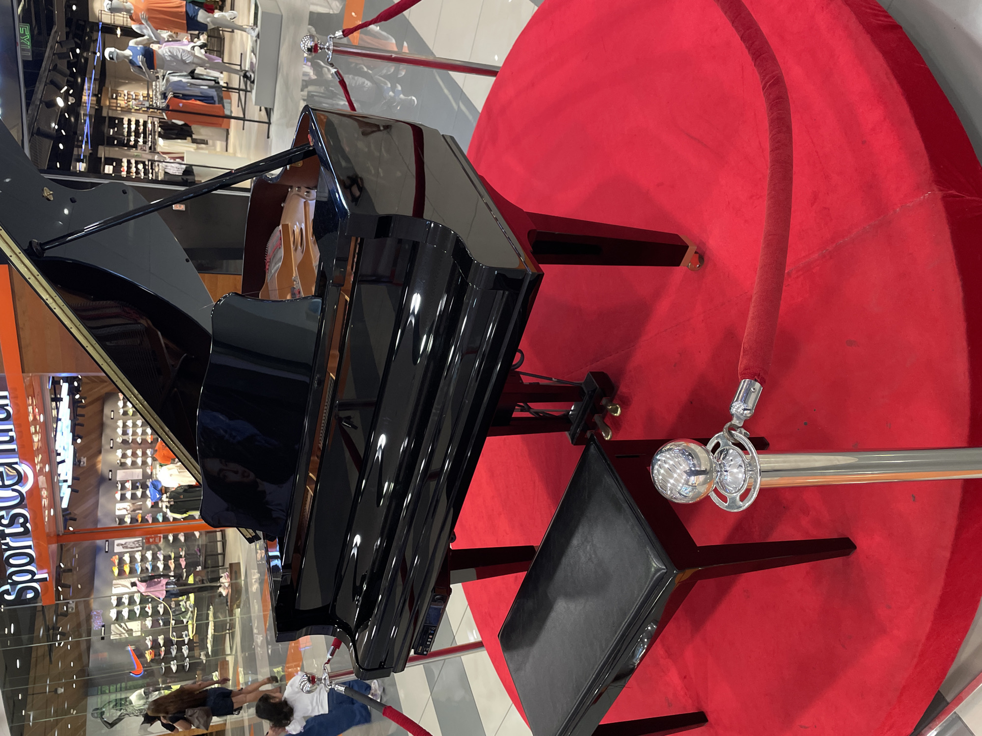 A glossy black grand piano is the central focus, positioned on a large, circular red carpet or platform. A matching black piano bench sits in front of the instrument, which has its lid open. The display area is cordoned off by several chrome stanchions with red velvet ropes.
In the background, a vibrant shopping mall environment is visible, featuring multiple retail stores with glass facades. Mannequins, dressed in various clothing items including athletic wear, are displayed inside these stores. Racks of clothing and shelves filled with shoes are also noticeable. The words "Sports Center" are partially visible on a store sign, and an "EXIT" sign can be seen further back. The polished surfaces of the piano reflect the overhead lights and parts of the surrounding mall. The floor is made of light-colored tiles. A few shoppers are present in the background, with one person appearing to lean against a stanchion near the piano display. The setting suggests an indoor commercial space, likely a mall, during daytime hours, with the piano set up as a display or for an event.