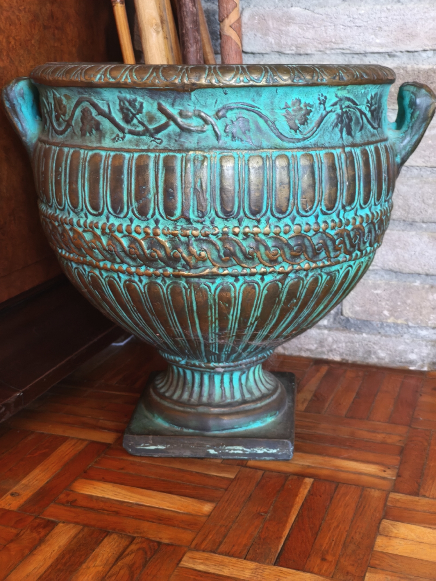A tall, ornate urn with a weathered, verdigris patina sits on a dark wooden floor with a parquet pattern. The urn has decorative fluting and a band of raised ornamentation around its midsection, featuring a vine motif. The background shows a stone wall and a dark wooden surface. The scene is indoors and does not depict any specific identifiable landmarks of Passignano sul Trasimeno, Italy, and no people are present.