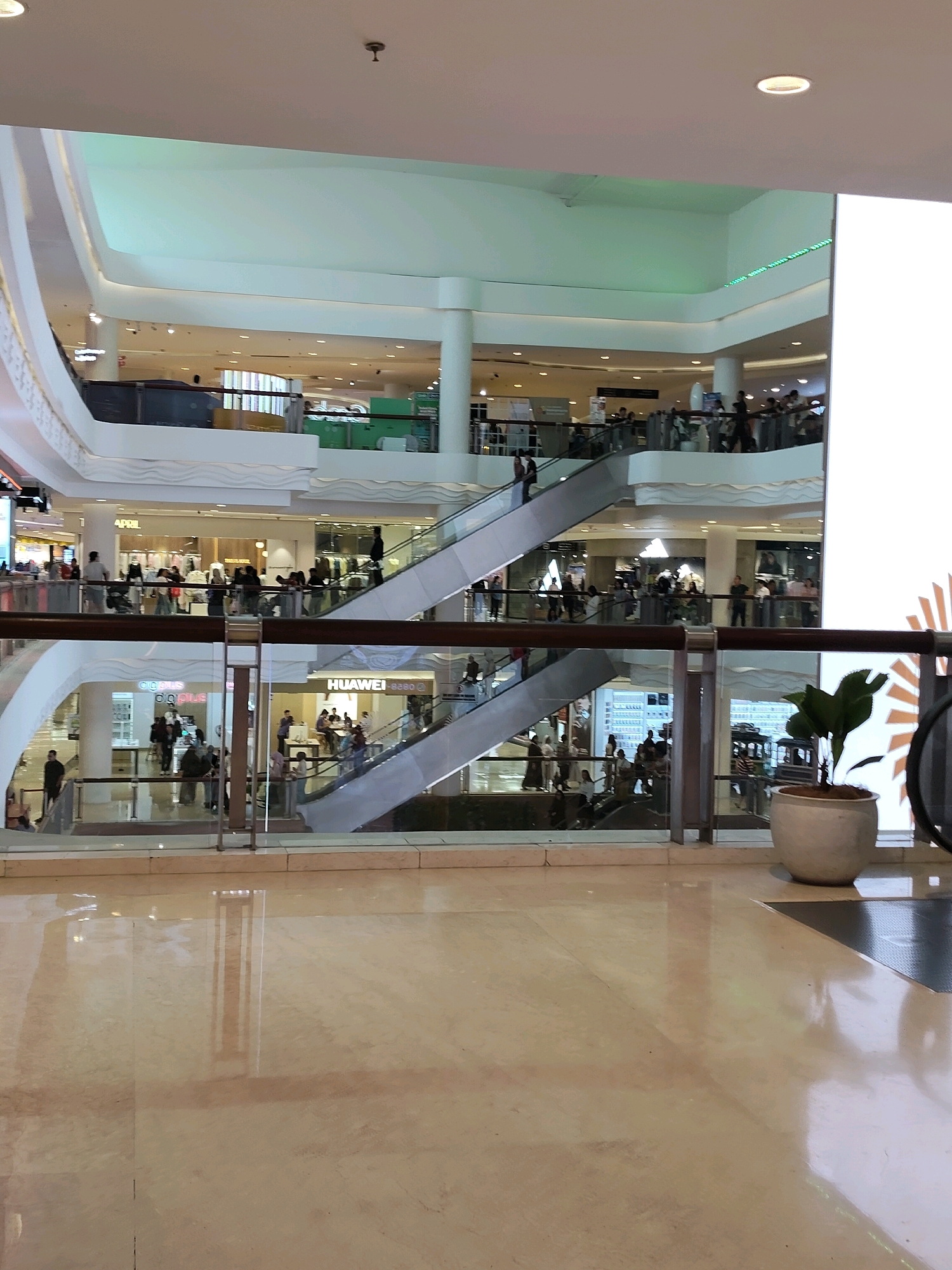 The image captures the interior of a multi-level shopping mall, likely located in the city of Medan, Indonesia, given the presence of a prominent "HUAWEI" store sign. The scene is brightly lit, reflecting off the polished marble floor. Escalators are visible, facilitating movement between floors, with numerous shoppers navigating the walkways and riding the escalators. Shops line the different levels, featuring signs like "APRIL" and "Cooplus." The architecture is modern, with curved white structures and ample glass railings providing visibility across the levels. In the foreground, a potted plant with large green leaves sits near a railing, adding a touch of nature to the commercial environment. The overall atmosphere suggests a busy and active shopping center.