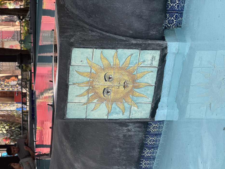 This image captures a section of a decorative fountain located in the Plaza de los Artesanos, Coyoacán, Mexico City, Mexico. The primary subject is a dark grey, textured concrete fountain wall rising from a light blue pool of water in the foreground. Centrally affixed to this wall is a square mosaic panel, composed of multiple light teal tiles, featuring a stylized yellow sun with a human face and radiating rays. Below this central mosaic, a band of smaller blue and white patterned tiles lines the fountain's edge at the water level. The calm water reflects a distorted image of the sun mosaic, alongside prominent streaks of vibrant red and pink hues originating from unseen structures or lighting in the background. Two individuals are partially visible in the upper-left background, positioned on the far side of the fountain. One male, wearing a grey shirt, is observing a mobile phone, while a female in a light grey top stands adjacent to him. Blurred architectural elements and faint greenery are discernible in the distant background.