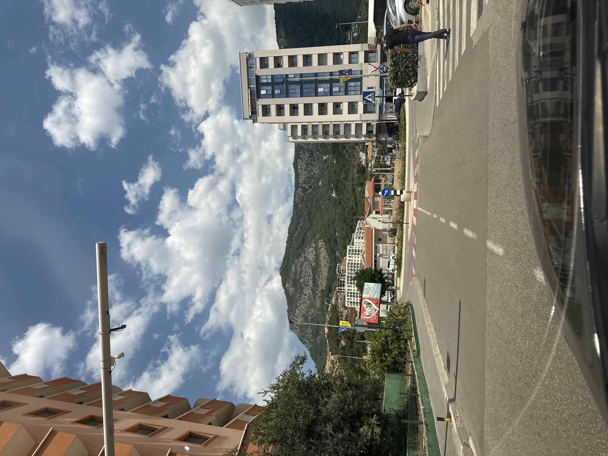 The image depicts a street scene in Budva, Montenegro, on a sunny day with scattered clouds. In the foreground, the hood of a dark car is visible, reflecting buildings and the sky. The street is paved with asphalt, marked with white lines. A pedestrian crossing is present on the right side of the road.
To the right, a tall, modern building with multiple floors and balconies stands prominently. It is a light-colored structure with many windows. A person wearing a black t-shirt and jeans is walking across the pedestrian crossing, away from the viewer. Further down the street, other buildings of varying architectural styles are visible, including lower-rise structures with red tile roofs.
On the left side of the street, a multi-story building with an orange facade and balconies occupies a significant portion of the frame. Adjacent to it is a green fence and some vegetation. In the background, a large, tree-covered mountain dominates the landscape, with some buildings nestled on its lower slopes. There are also streetlights and signs present. A billboard with an advertisement is visible in the middle ground. The overall impression is of a developed urban area with a backdrop of natural scenery.