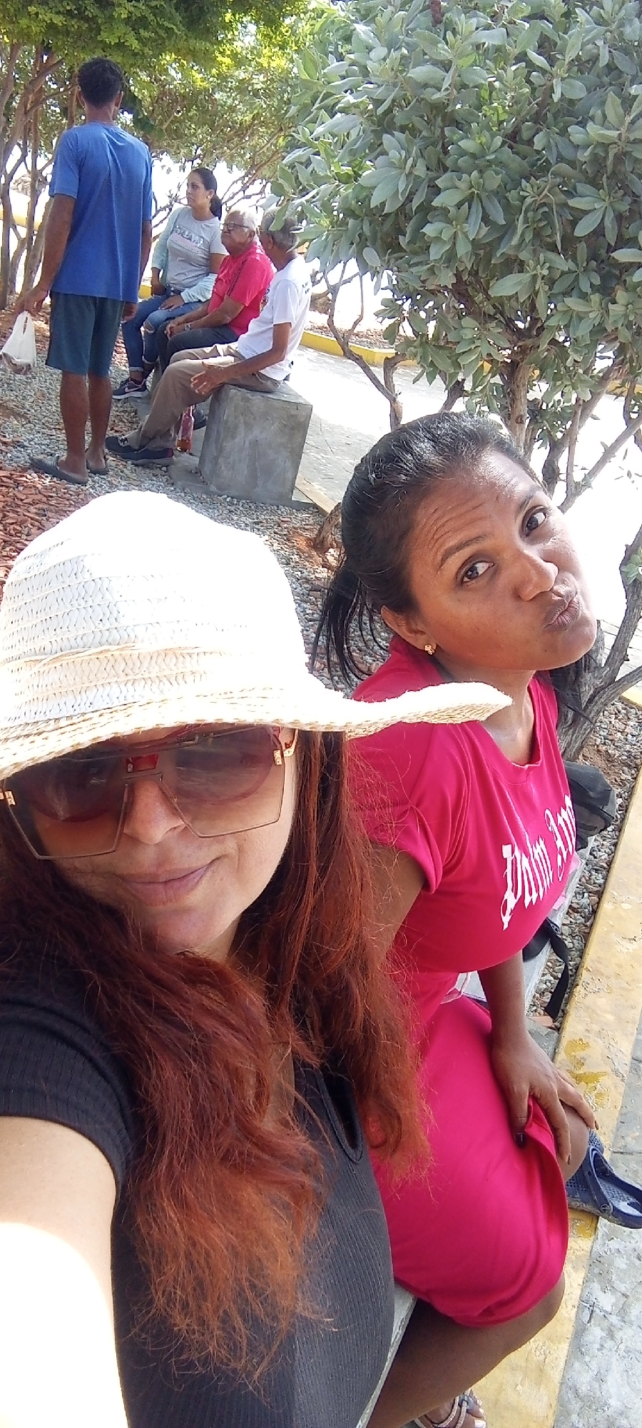 The image captures a scene in an outdoor location. In the foreground, two women pose for a selfie. The woman on the left wears a wide-brimmed straw hat and sunglasses, with red hair. The woman on the right is wearing a pink t-shirt and making a kissing face towards the camera.
In the background, a group of people are seated on benches and concrete blocks. One man is standing, wearing a blue t-shirt and shorts, and carrying a plastic bag. The environment includes trees and what appears to be a gravel or pebble-strewn ground. In the far background is a body of water.
The location is in the city of Unknown, Unknown.