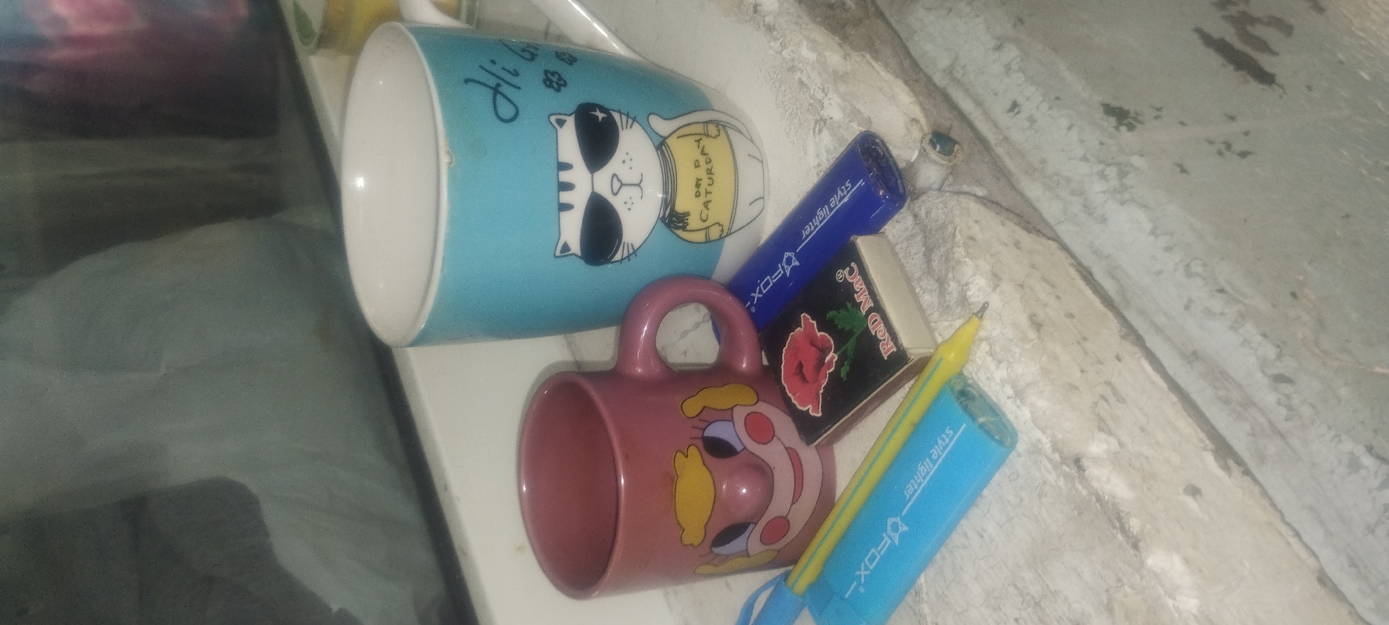 The image shows a still life arrangement of two mugs and a few small objects, likely on a windowsill or shelf. In the foreground, a pink mug with a cartoonish face is positioned on its side, revealing its interior. Behind it, a larger, light blue mug is also tilted, displaying a graphic of a cat wearing sunglasses and a shirt that reads "DAY P CATURDAY". The cat is drawn with a playful expression.
Scattered around the mugs are several small items. A blue "Style Lighter" with a "FOX" logo is visible, as is a yellow pencil. A small black box with a red rose graphic and the text "Red Mac" is also present. These items suggest a casual, possibly domestic setting. The overall lighting is dim, suggesting it could be evening or indoors with artificial light. The background appears to be a textured, possibly concrete or stone surface, common in many Ukrainian locations, including Zaporizhzhia. There are no people in the image, and no discernible weather conditions. The arrangement of objects implies a moment of stillness, with no active event taking place.