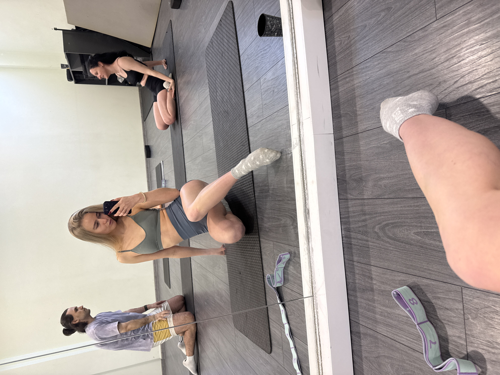 A mirror selfie in a fitness studio with multiple people in the reflection. The foreground has a partial view of a person's leg with a sock. A gray stretching band rests on the gray wood-like floor near the mirror. The mirror reflects a woman with long blonde hair taking the selfie. She is wearing a gray sports bra and shorts while kneeling on a black yoga mat. Another woman with long dark hair kneels on a yoga mat in the background. A man with a bun and purple shirt sits in the lower left corner. A black storage unit is in the upper left background.