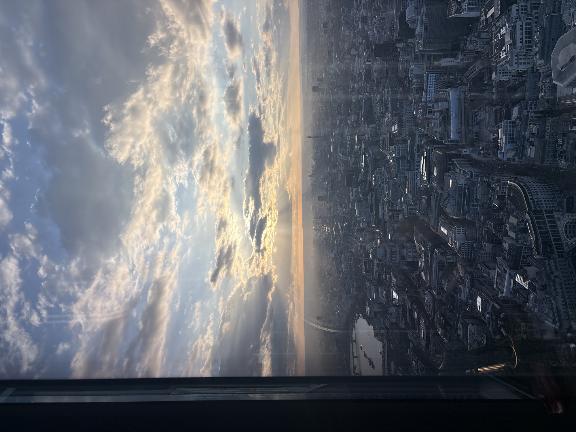 The image is an aerial view of London, United Kingdom, taken from a high vantage point, possibly from a tall building or observation deck. The city is split in half. The left side is the sky, filled with clouds illuminated by the setting sun, creating a warm, golden light. The right side is the city, filled with buildings and infrastructure. It also includes a large river with a bridge across it. In the distance, a tall, thin spire can be seen. There are no people visible. The image has been rotated 90 degrees counterclockwise.
