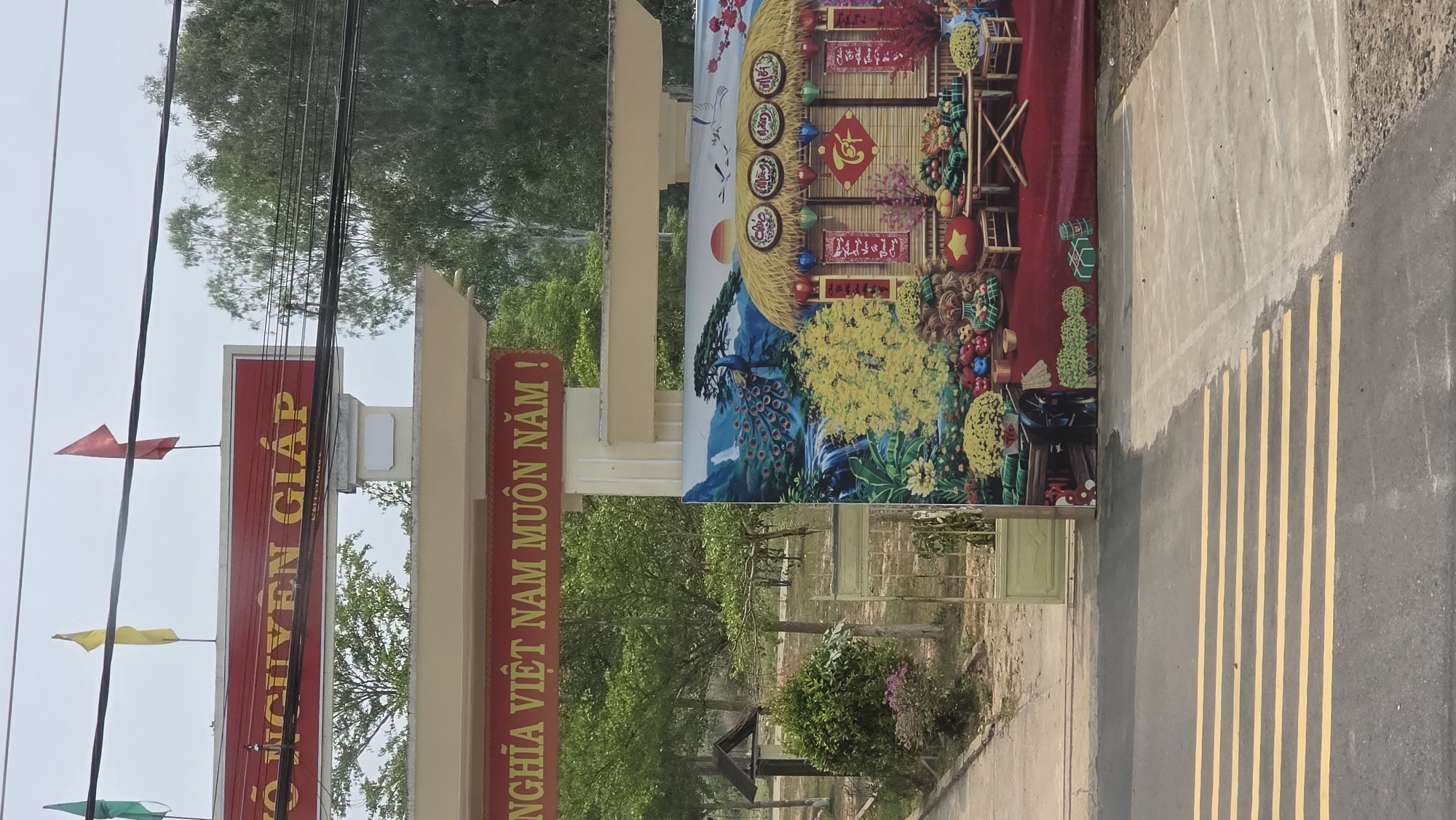 The image shows an entrance gate in what appears to be Đăk La, Vietnam. The gate is constructed of beige-colored concrete pillars and beams. On the left pillar, there's a long red sign with yellow Vietnamese script, topped with a small red flag. Other flags are present. The right pillar has a similar red sign, also with yellow Vietnamese script. Between the pillars, a large, colorful decorative panel is affixed. The panel depicts a traditional Vietnamese scene, featuring a house decorated for Tết (Lunar New Year), with images of flowers, fruits, a peacock, and other festive elements. The panel is set against a backdrop of what appears to be mountains and trees. Beyond the gate, a grassy area with sparse vegetation is visible. A paved road with yellow lane markings is in the foreground on the right. Overhead, there are several power lines. The sky is overcast.