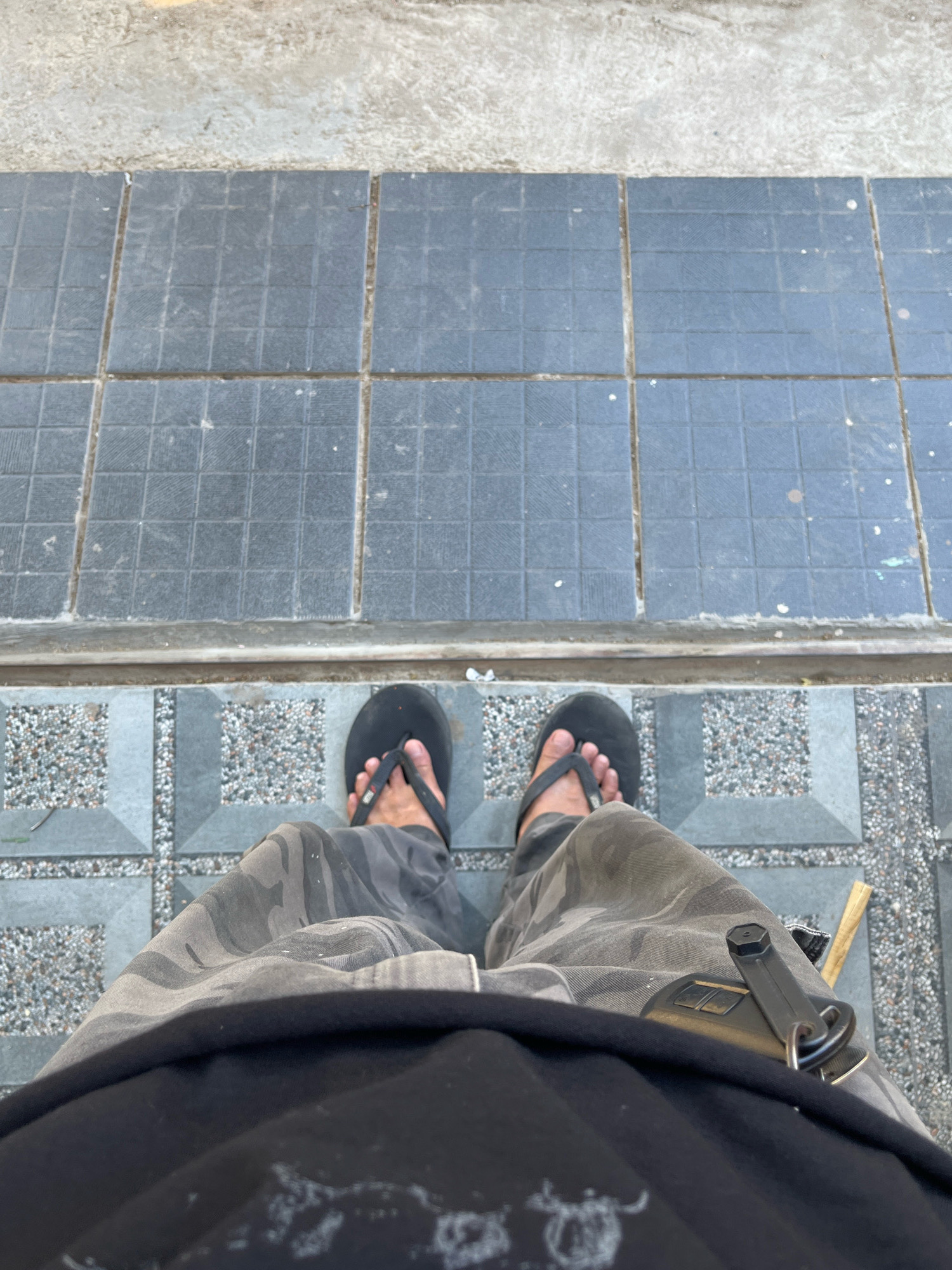 A first-person perspective shot shows a person wearing black flip-flops and camouflage pants. The person is standing on a tiled surface that transitions from a larger, dark gray tiled area to a smaller, patterned tiled area with gravel. A key fob is attached to the person's pants pocket, and a wooden object is visible to the right of the key fob. The scene appears to be outdoors, possibly on a walkway or patio. There is no information provided about the location in Tungkaran Pangeran, Indonesia.