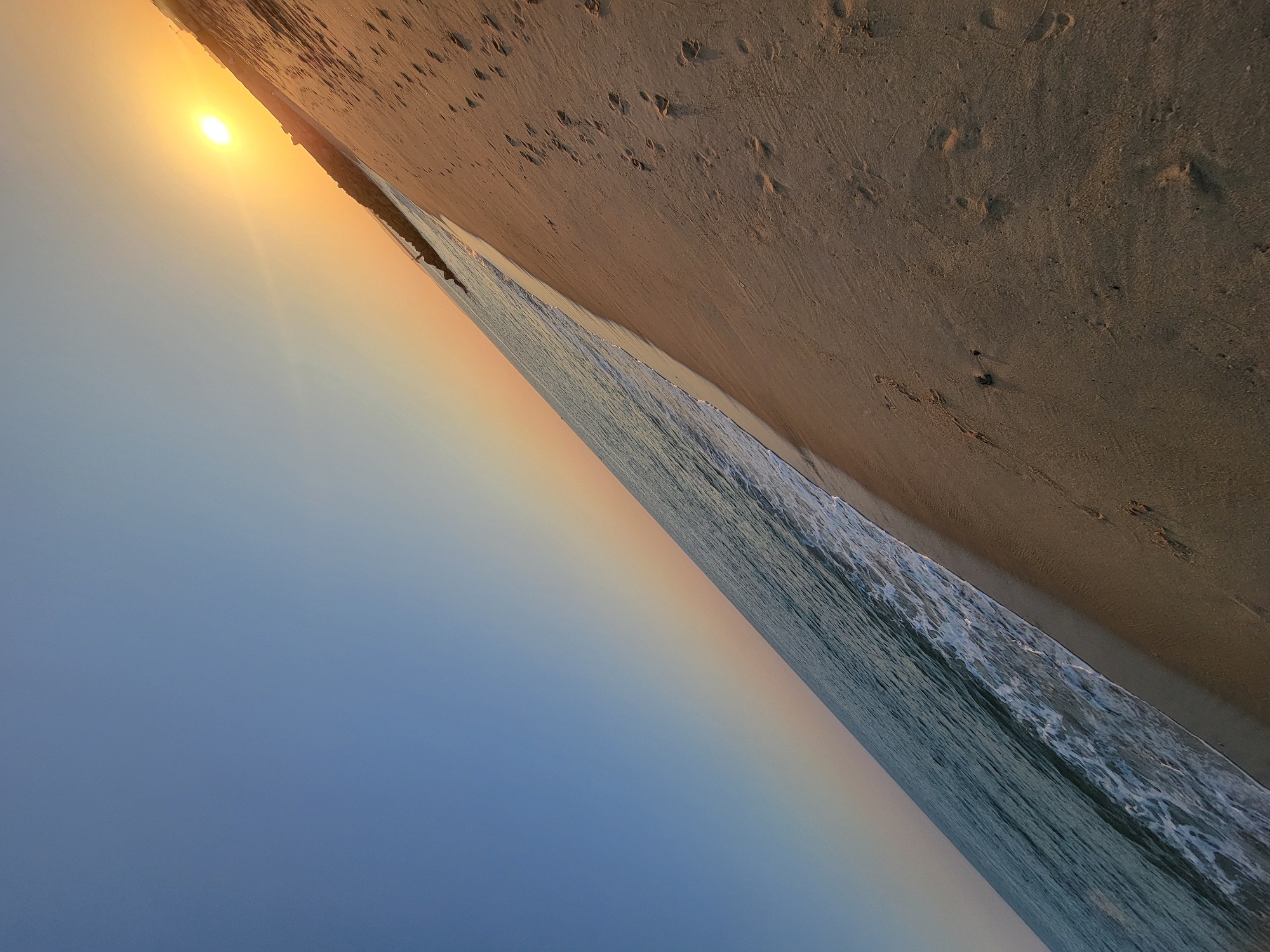 The image depicts a wide-angle view of a beach at sunset, likely in Quarteira, Portugal, due to the presence of the breakwater characteristic of the area. The scene is captured at an angle, with the horizon running diagonally across the frame. The sky transitions from a clear, deep blue at the top left to a soft gradient of orange and yellow near the setting sun, which is positioned in the upper right quadrant.
The ocean occupies the left side of the image, with gentle waves lapping at the shore, creating white foam. The water appears a muted blue-green. Adjacent to the water is a sandy beach, illuminated by the warm light of the setting sun. Footprints are visible in the sand, indicating recent human presence. In the distance, along the shoreline, a dark, linear structure, likely a breakwater or pier, is discernible, with a few scattered rocks. The overall impression is one of a tranquil end to the day at a coastal location.