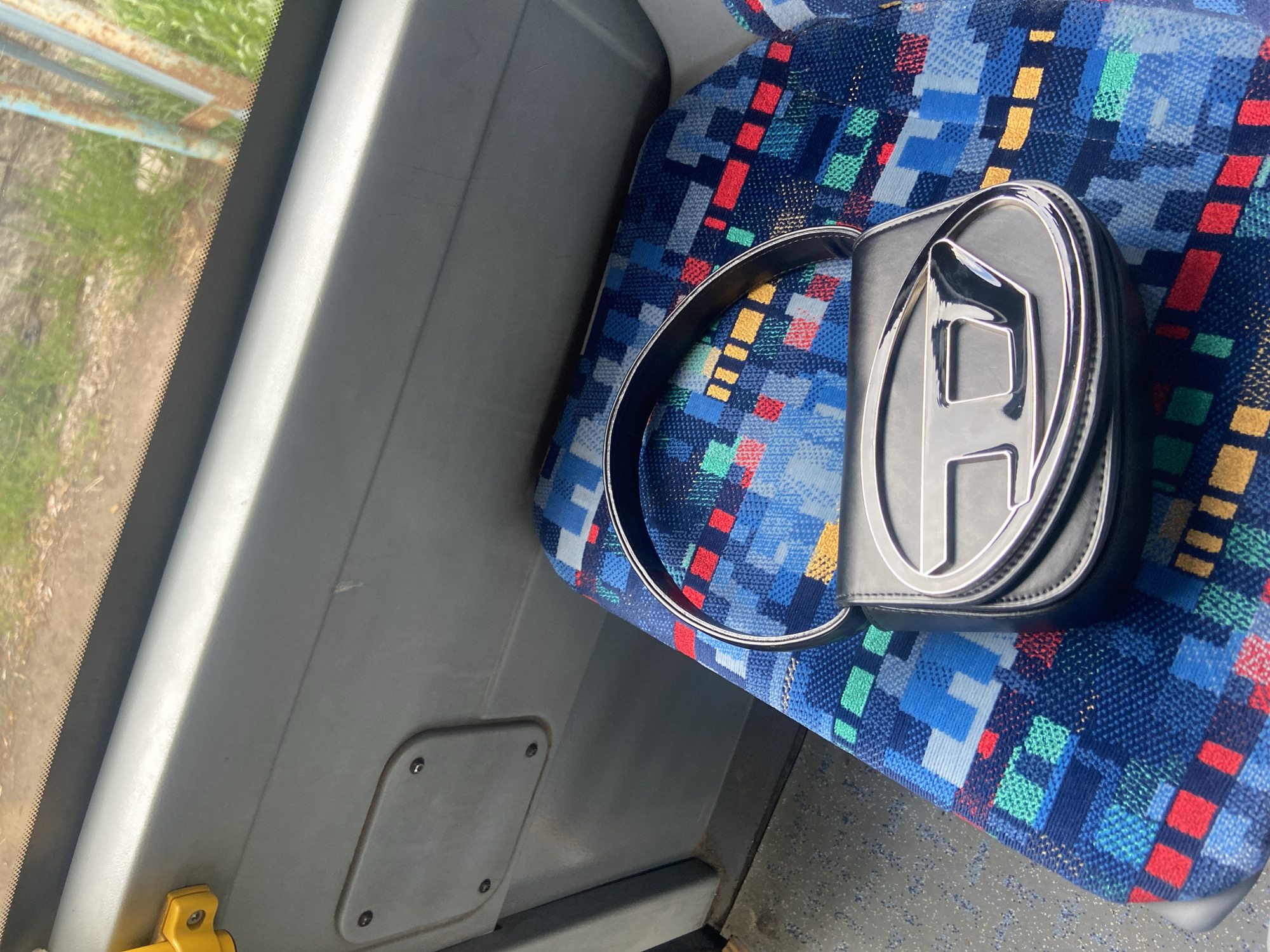 The image captures a scene inside a vehicle, likely a bus or coach, with a black bag resting on a patterned seat. The seat is upholstered in a vibrant, pixelated or geometric design featuring various shades of blue, red, green, and yellow. The bag, positioned towards the right of the frame, has a prominent, glossy silver metallic logo on its flap, resembling a stylized letter 'D', consistent with the Diesel brand.
Through the window on the left, a bright outdoor scene is visible, dominated by green grass and patches of dirt, with a rusty, blue-tinged metal fence or gate structure in the background, suggesting a rural or semi-rural environment. The window has a visible rubber seal along its edge. Light gray interior paneling of the vehicle runs along the window and beneath the seat, and a small yellow handle or latch is partially visible in the bottom left corner. The vehicle's floor, with a speckled gray pattern, is visible in the bottom right.
The scene depicts a static moment, with no people visible. The bright lighting indicates it is daytime, likely sunny or partly sunny. The setting suggests a journey through Zhiten, Bulgaria.