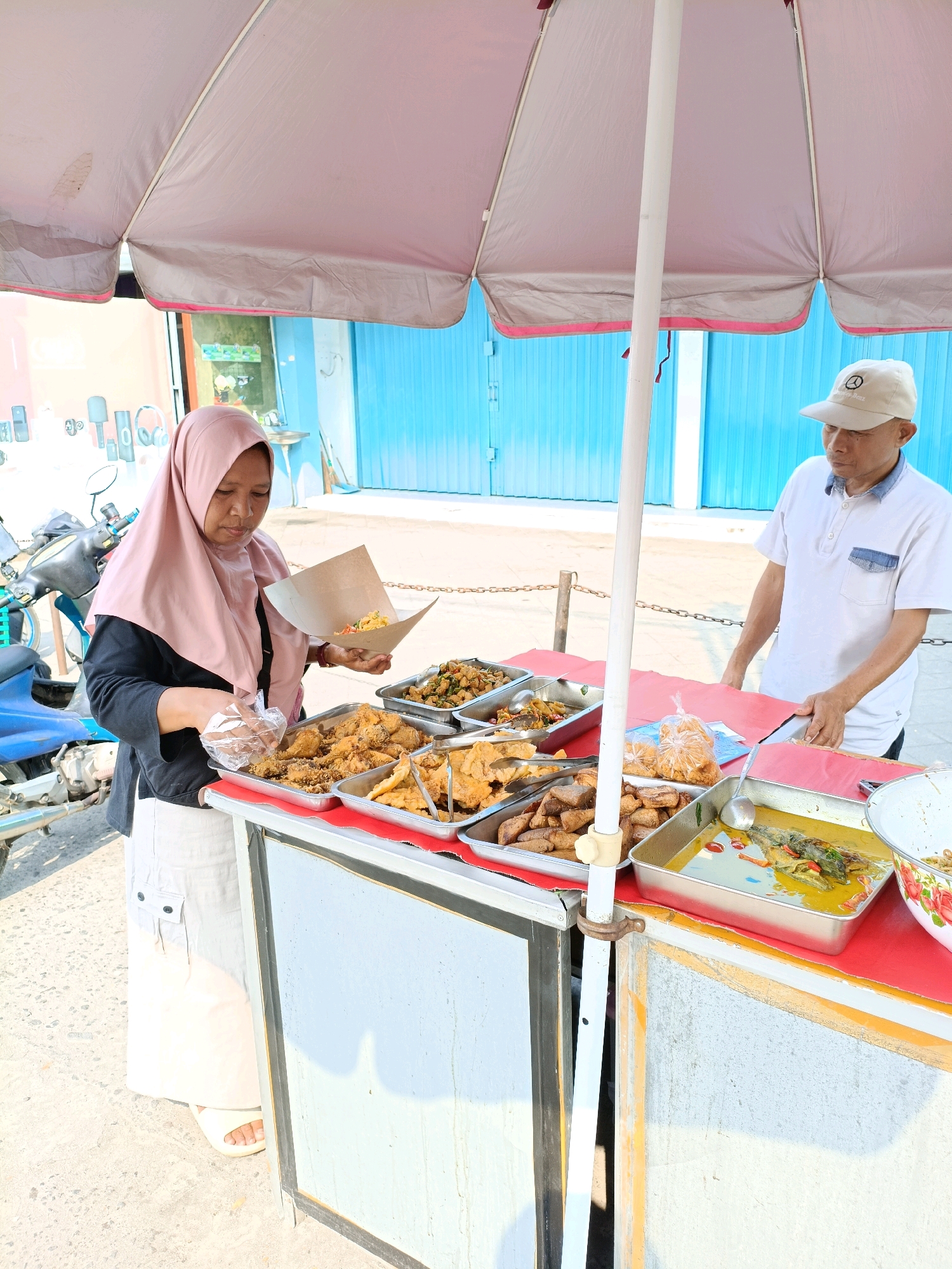 In Maguwoharjo, Indonesia, a street food vendor, a woman wearing a pink hijab and a black shirt, is serving food from a cart. She is holding a paper cone filled with food and a plastic bag in her hands, appearing to be in the process of portioning it. In front of her, on the cart's red-covered surface, are several metal trays containing various prepared dishes, including what appears to be fried chicken, tofu, and a yellow curry with fish. A man in a white polo shirt and a beige cap stands to the right, assisting at the cart. The scene is outdoors, with a bright blue corrugated metal wall visible in the background and a motorcycle parked to the left. The overall environment suggests a sunny day at a local market or roadside stall.