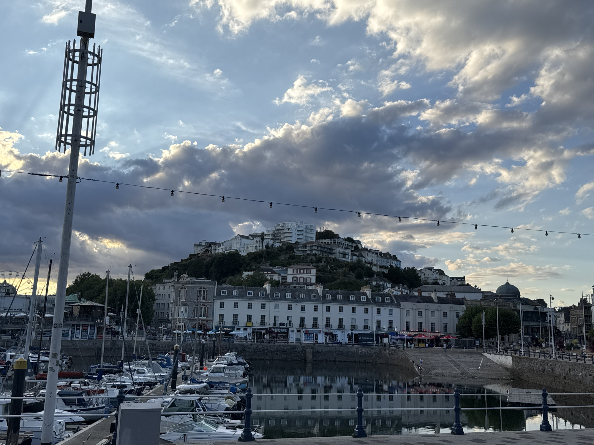 This image captures a scenic view of Torquay harbor, United Kingdom, during what appears to be late afternoon or early evening. The foreground is dominated by a marina filled with numerous sailboats and smaller motorboats docked in calm water. A prominent light pole with a cage-like structure and a small box at its top stands to the left. A string of decorative lights stretches across the frame, low in the sky.
Beyond the harbor, a row of white buildings with multiple windows and dark roofs lines the waterfront. Behind these buildings, the town of Torquay rises up a steep hill, with a dense collection of residential buildings and some larger structures, including a white, multi-story building at the peak. The architecture suggests a mix of historical and modern styles. Lush green trees are interspersed among the buildings on the hillside.
The sky is dramatic, with a mix of dark, heavy clouds and patches of bright blue sky, indicating dynamic weather conditions. Sunlight is breaking through some of the clouds, casting rays onto the scene. The water in the harbor reflects the buildings and the sky, creating a mirror-like effect. There are a few people visible on the waterfront promenade and near some of the buildings, and possibly in the boats. In the distance, to the left, a Ferris wheel can be seen, suggesting an amusement area or fairground. The overall atmosphere is serene yet visually striking due to the interplay of light and shadow.