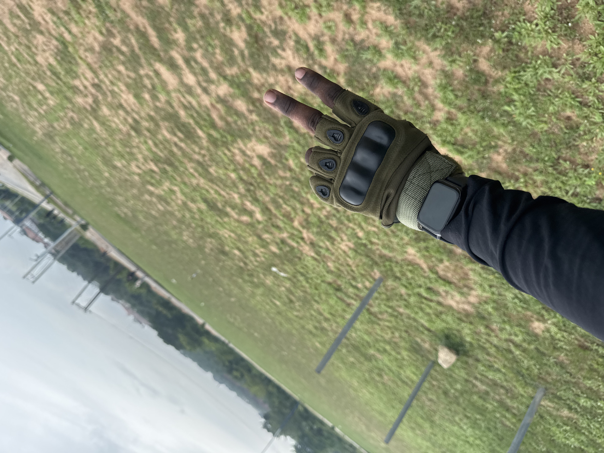 The image shows a first-person view of a person's arm and hand making a peace sign. The person is wearing a tactical glove and a smartwatch. The background consists of a grassy field and what appears to be a body of water or swamp. The field has visible support poles in several places. Based on the context provided, the location is likely Ihiagwa, Nigeria.