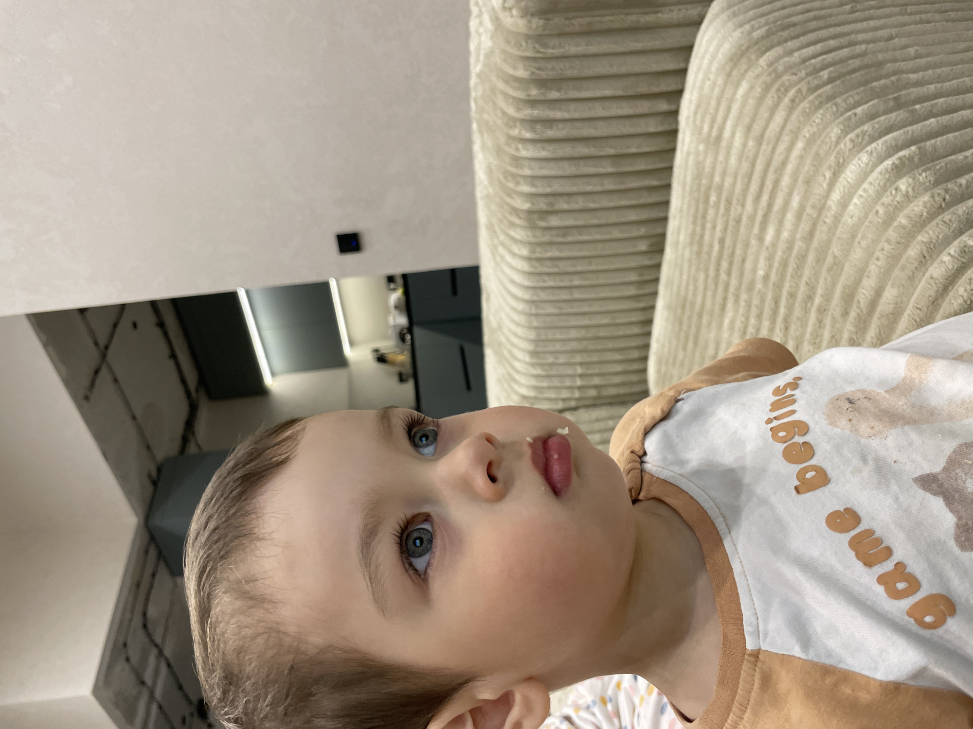 The image captures a close-up portrait of a young child, likely a toddler, with light brown hair and striking blue eyes. The child is positioned in the foreground, looking intently upwards and slightly to the right. A small, light-colored crumb or piece of food is visible on their lower lip. They are wearing a two-toned t-shirt: a white central panel combined with brown sleeves and collar. On the white section, the words "Game Begins" are visible in raised orange lettering, alongside some lighter brown, abstract shapes which might be part of an animal design.
To the right of the child, a large section of a light beige or cream-colored, ribbed sofa or armchair is prominent, adding a soft, textured element to the composition. The background features a plain, light-colored wall or ceiling. Below this, reflections from modern, dark-toned surfaces, possibly kitchen cabinets or sleek furniture, are visible. A small dark square object with a blue indicator light can also be seen on the wall in the upper-middle background. The scene suggests an indoor setting, likely a home, located in Izhevsk, Russia. The child's expression appears calm and observant.