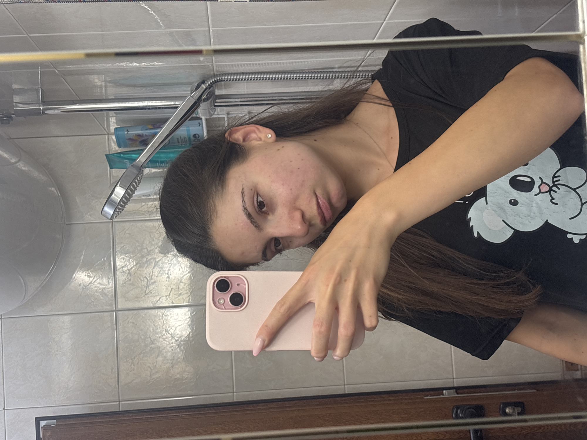 The media file is a mirror selfie taken by a young woman in a bathroom. The woman has long, dark brown hair, and is wearing a black t-shirt with a koala graphic on it. She is holding a light pink smartphone in her right hand and is looking at the camera. Her left arm is resting on her chest. The bathroom has tiled walls and a wooden cabinet is visible at the bottom of the frame. A shower head is also visible. The image appears to have been taken indoors, likely in a residential bathroom, given the presence of toiletries and the overall ambiance. The lighting suggests it could be daytime, but without further context, the exact time of day is uncertain. The woman's expression is neutral. The location is identified as Varna, Bulgaria. There is no visible text in the image.