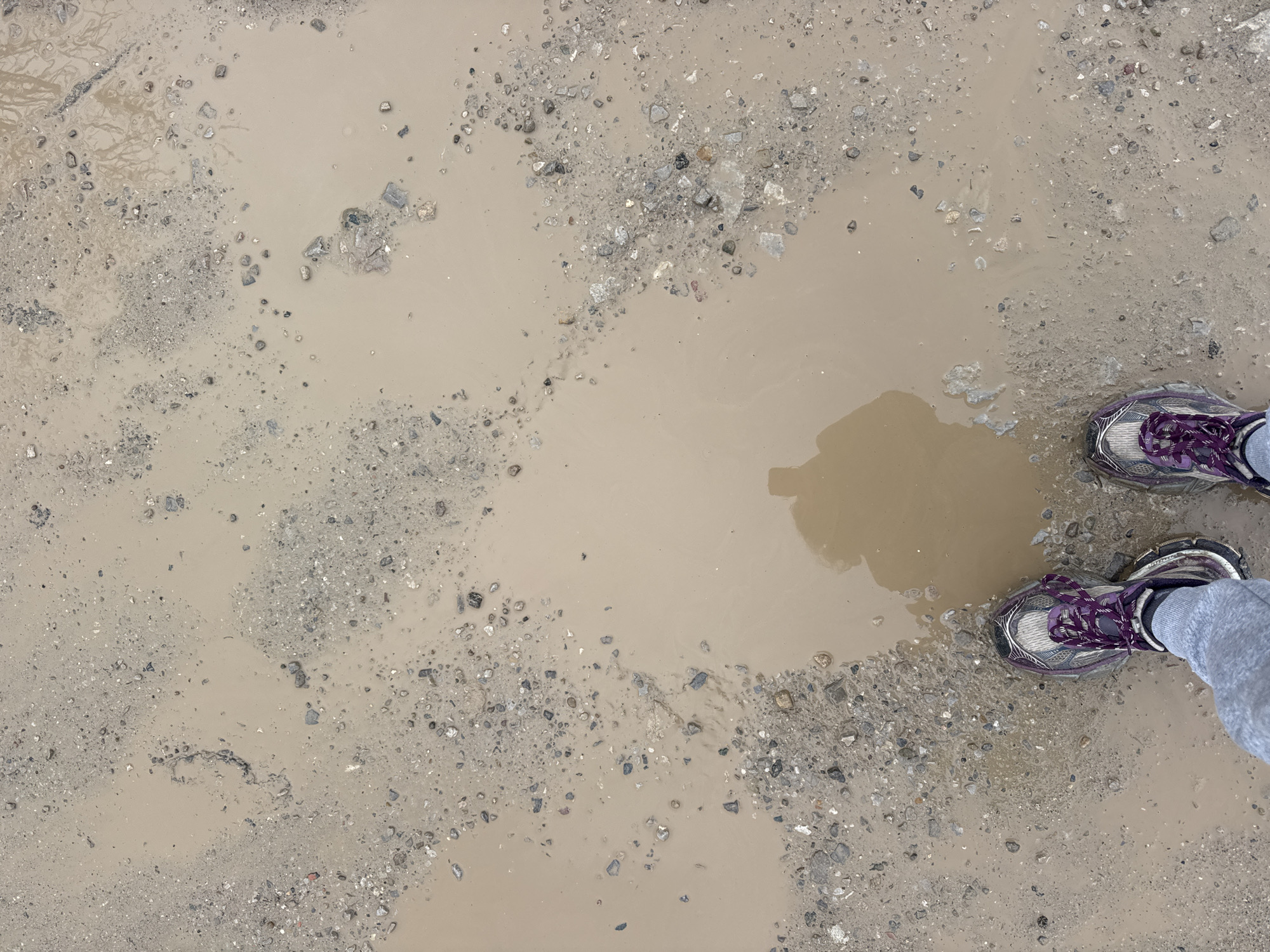 An eye-level, medium-angle shot depicts a gravelly, muddy surface with a large puddle. A person, visible only from the knees down, stands on the edge of the puddle. The person is wearing gray sweatpants and purple and gray athletic shoes. The puddle reflects a silhouette. The scene implies an outdoor environment with wet or recently wet conditions. This location cannot be determined to be in Sofia, Bulgaria based on the available information.