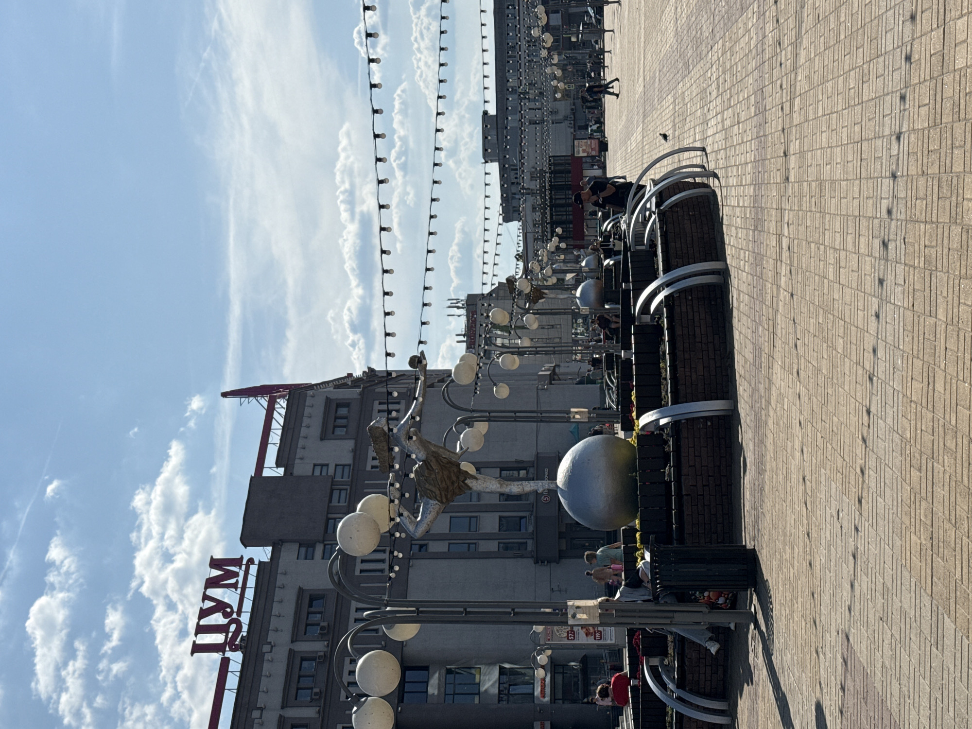 The image depicts a street scene in Nizhny Novgorod, Russia, likely on Bolshaya Pokrovskaya Street. In the foreground, a paved pedestrian area with a brick pattern is visible, with several dark trash bins and benches placed along a raised flower bed. A sculpture of a dancer on a sphere stands prominently in the mid-ground, surrounded by decorative lamps and more flower beds. In the background, a large building with the red Cyrillic lettering "ЦУМ" (TSUM) is visible, indicating a department store. Several people are present in the scene, some sitting on benches and others walking. The sky is blue with scattered white clouds, suggesting a daytime setting. String lights are hung across the street above the pedestrian area.
