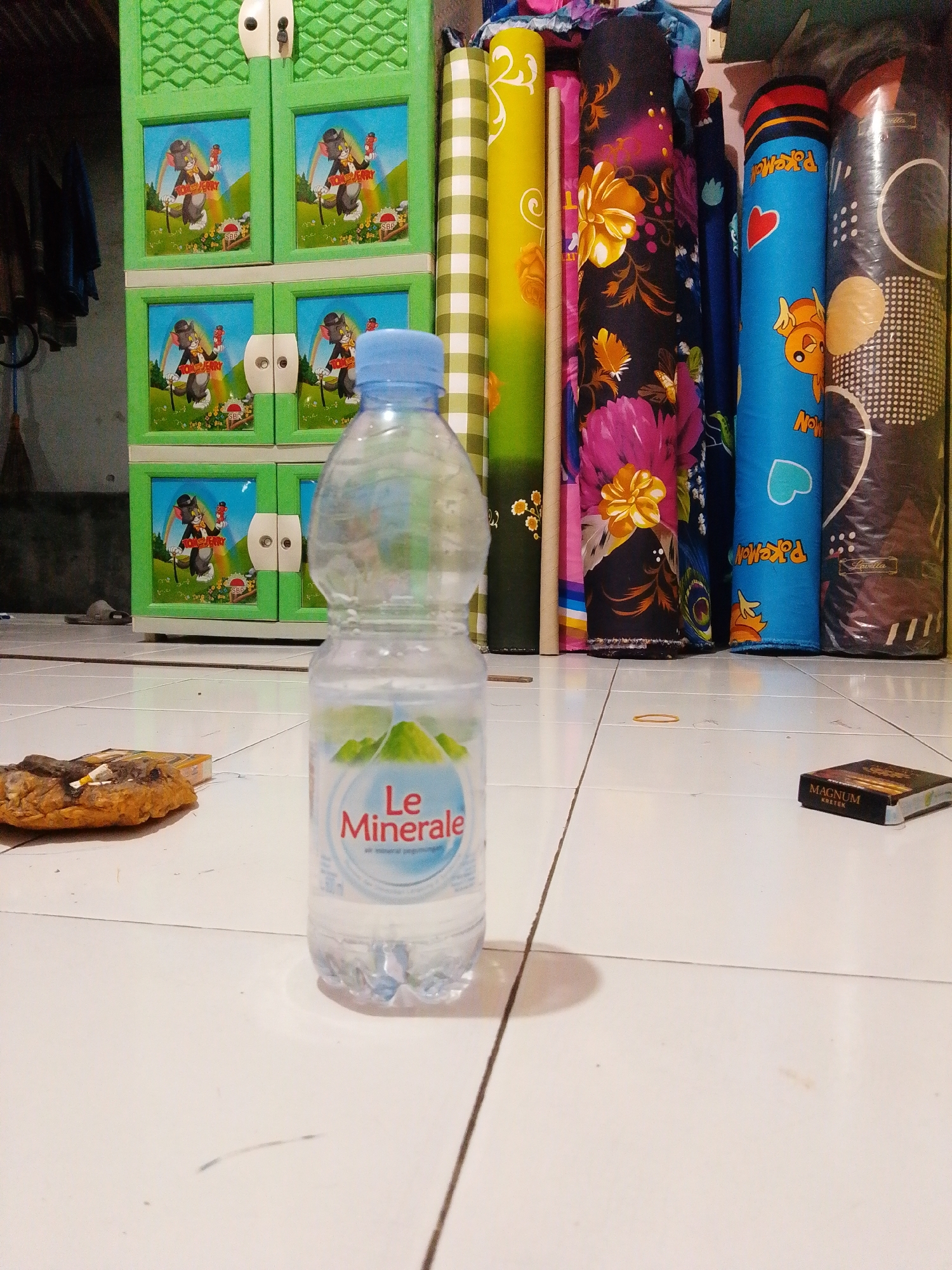 In Bohar, Indonesia, a low-angle shot captures a scene on a tiled floor. In the foreground, a clear plastic bottle of Le Minerale water stands upright. To its left, a crumpled brown object, possibly food waste, has cigarette butts placed on it. To the right of the water bottle, a pack of Magnum Kretek cigarettes rests on the floor. In the background, a green plastic cabinet with four illustrated doors featuring Tom and Jerry characters is visible. Adjacent to the cabinet are several rolled-up fabric items with various colorful patterns, including one with a checkered green and white design and another with floral motifs. The lighting suggests an indoor environment, possibly a room in a residential or commercial space in Bohar.