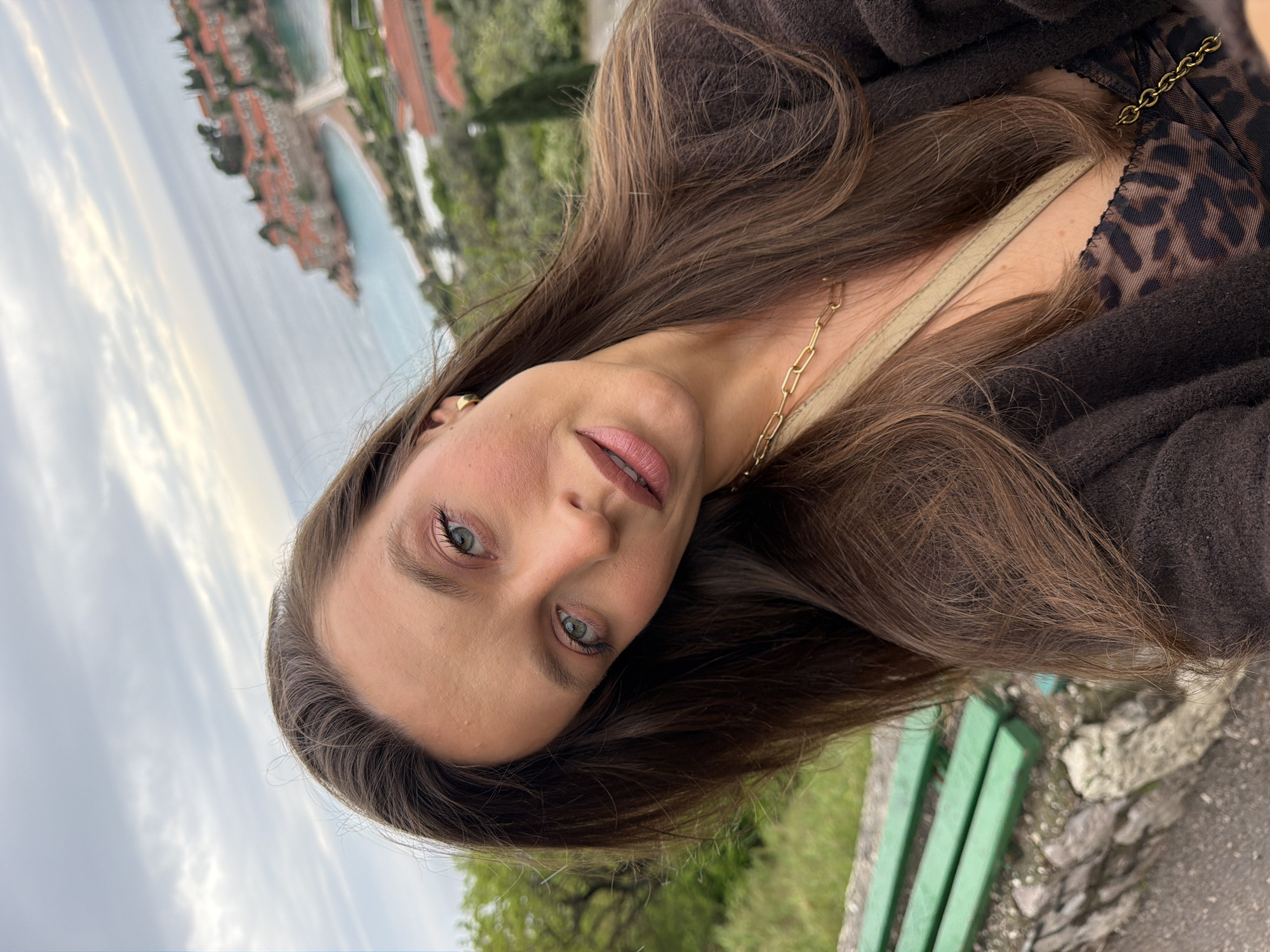 A medium-close up portrait features a young woman with long, straight, medium-brown hair, looking directly at the camera with a calm and direct expression. Her head is tilted, revealing her striking light green/hazel eyes, light pink/mauve lips, and natural-looking makeup with a touch of blush. She wears small gold hoop earrings and a gold chain necklace. Her attire includes a dark brown, possibly knitted, outer layer, worn over a brown and black leopard print top. A light beige strap, likely from a bag, is visible over her shoulder.
The background presents a scenic coastal landscape, identifiable as Sveti Stefan, Montenegro. A cluster of red-roofed buildings, characteristic of the fortified island village, is visible on a landmass surrounded by water. Abundant green foliage covers the land, and the sky above is overcast with soft clouds, indicating daylight. In the bottom right foreground, a part of a green slatted bench and some rocky ground are visible. The image is captured from a slightly tilted perspective. There is no visible text in the image.