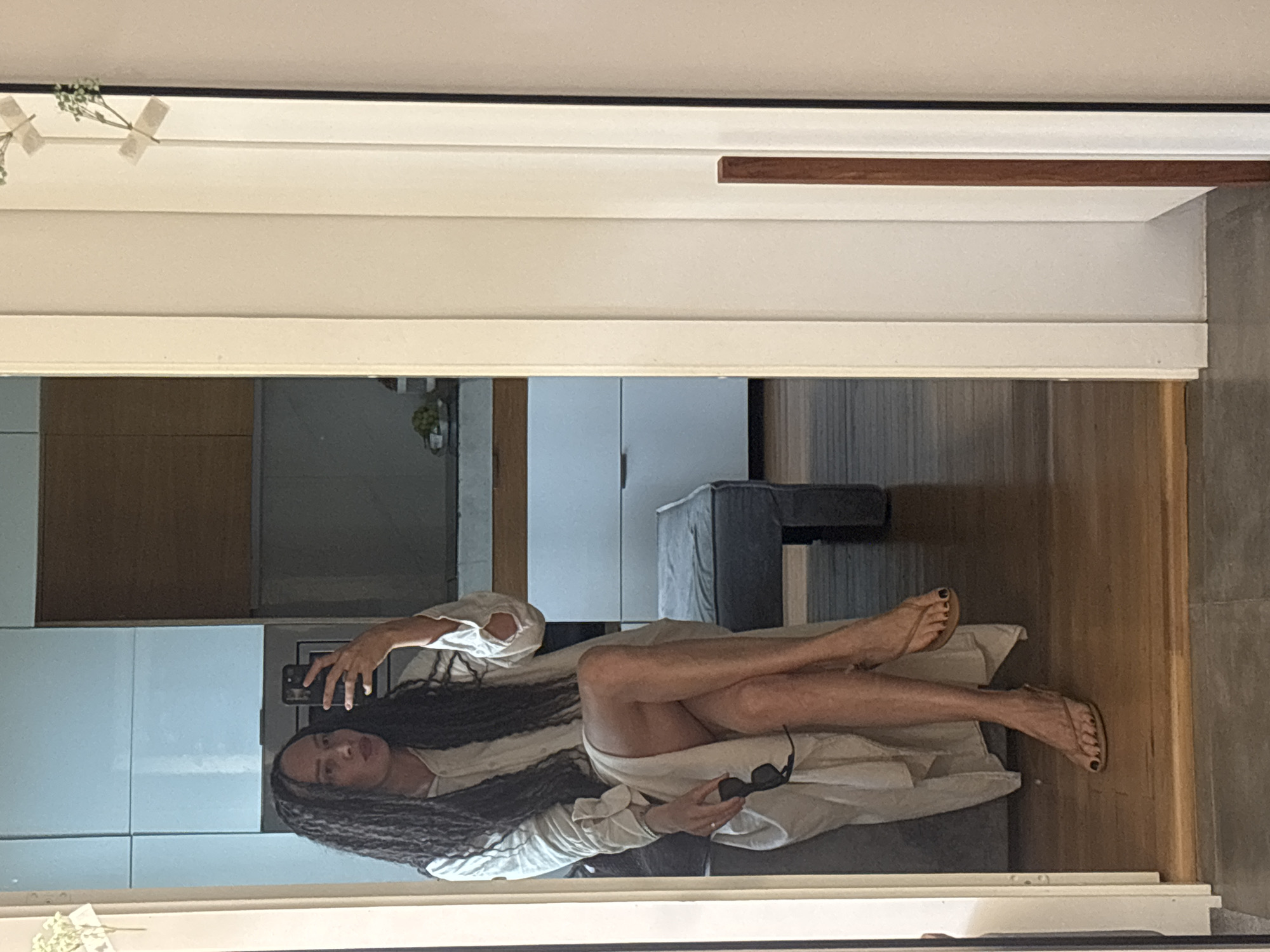 A full-length reflection of a woman taking a selfie in a mirror. The woman has long, dark dreadlocks and is wearing a light-colored dress or robe. She is holding a smartphone in her right hand, which is raised in front of her. In her left hand, she is holding sunglasses. The background of the reflection includes a kitchen area with light-colored cabinets and a gray couch. The environment appears to be a modern apartment. The location in the city of Chaikovsky, Russia is not verifiable from the image.