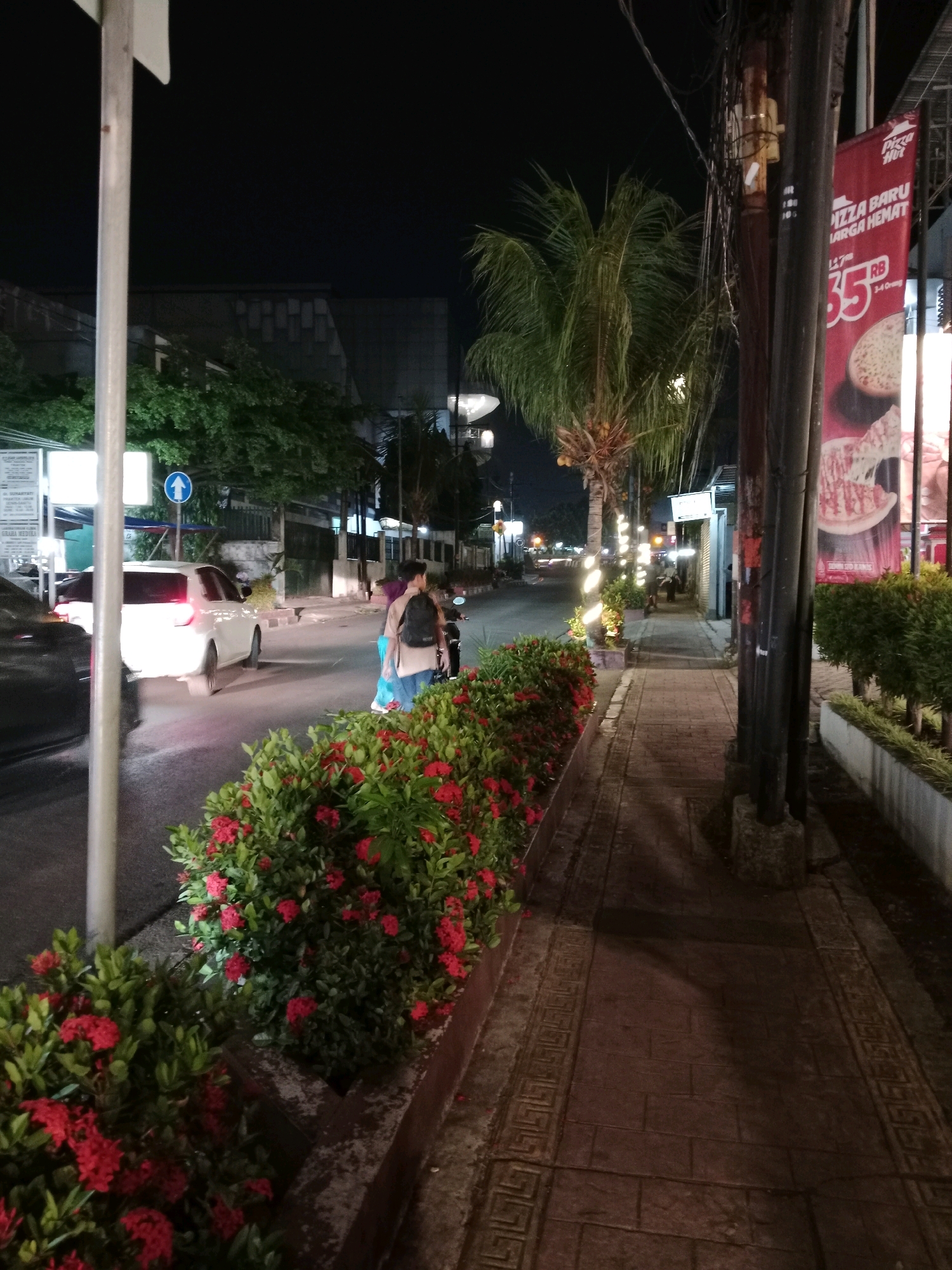 The image depicts a nighttime street scene in Cianjur, Indonesia. A sidewalk runs along the right side of the frame, paved with brick-like stones and adorned with a decorative border. A row of vibrant red ixora bushes lines the edge of the sidewalk, separating it from the street. On the street, a white car is visible, its taillights illuminated, driving away from the camera. To the left, a pedestrian signal with an upward-pointing blue arrow is mounted on a pole.
Further down the street, palm trees are illuminated, and distant traffic lights create a blurred effect. A Pizza Hut banner is prominently displayed on the right side of the image, advertising a new pizza and a discounted price of "35 RB". A dark-haired individual wearing a light brown shirt and a black backpack stands on the sidewalk near the red bushes, facing away from the camera. Another person, partially obscured, is visible behind them. Electrical poles and wires are scattered throughout the scene, common elements of urban infrastructure. The overall impression is of a lively, but dimly lit, street at night.