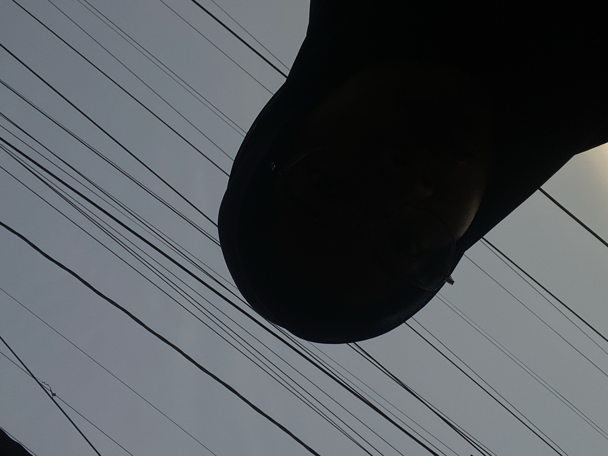 The image, taken from a low-angle perspective in Air Molek, Indonesia, shows a person wearing a dark hijab silhouetted against a pale sky crisscrossed with numerous black electrical wires. The wires are arranged diagonally, creating a sense of depth and complexity against the overcast sky. The subject's face is mostly obscured by shadow, with only the glint of glasses visible. The composition is dominated by the interplay of light and shadow, with the dark silhouette of the hijab and the stark lines of the wires contrasting sharply with the muted tones of the sky. The overall impression is one of urban anonymity and the ubiquity of electrical infrastructure in the cityscape.