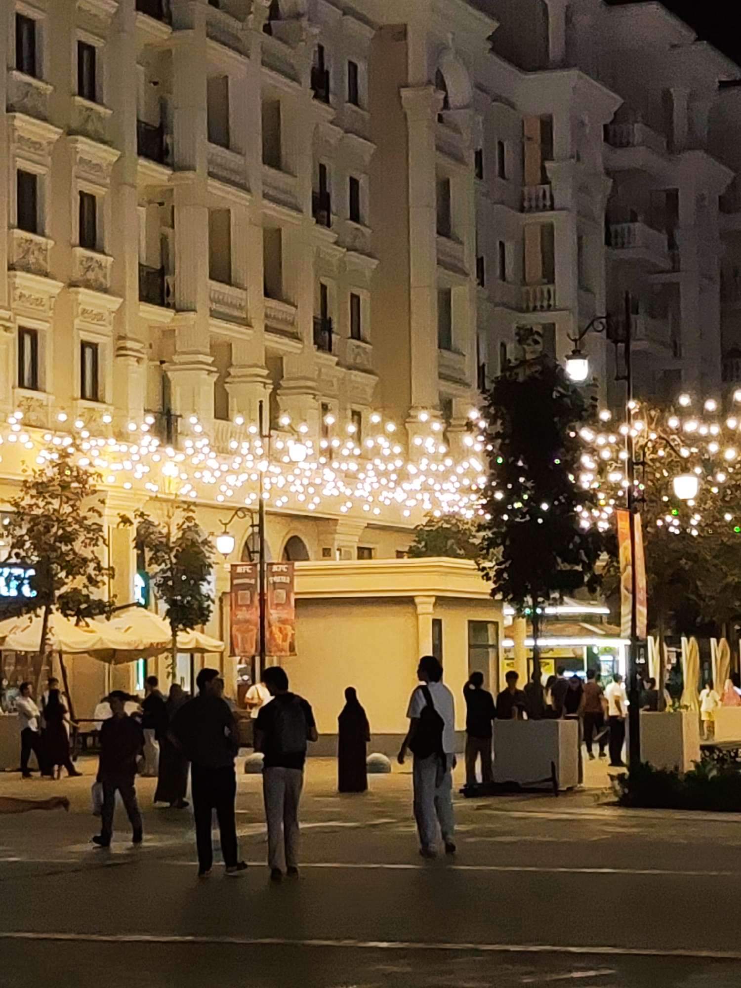 The image depicts a nighttime street scene in Tashkent, Uzbekistan, likely a pedestrianized area or a promenade. The architecture suggests a modern, upscale district, with a grand, multi-story building featuring intricate decorative elements, balconies, and numerous windows, all illuminated by warm, ambient light. String lights are draped across the street, creating a festive atmosphere, and are also visible on trees.
Several individuals are scattered across the paved area. In the foreground, a person wearing a white shirt and dark pants, with a backpack, walks away from the camera. To their right, another person stands with their back to the camera, wearing a dark shirt and light-colored pants. Further back, a group of people are gathered, some appearing to be in conversation. To the far left, a few individuals are near some white canopied structures, possibly outdoor seating for a cafe or restaurant. Two tall, red banners with text are visible hanging from a lamppost, partially obscured by trees and string lights. The overall impression is one of a lively urban space at night, with a mix of modern architecture and illuminated decorations.
