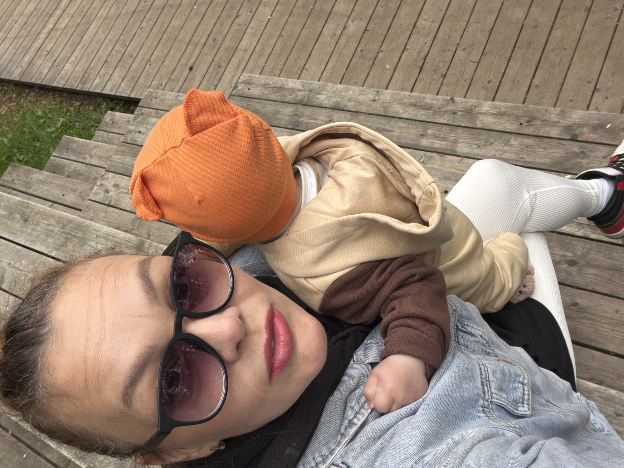 The media file shows a close-up, high-angle shot of a woman and a baby lying on a wooden outdoor staircase. The woman is in the foreground, facing upwards towards the camera. She is wearing sunglasses with large, dark frames and has on a light blue denim jacket and a dark scarf. Her lips are painted a bright pink. The baby is nestled beside her, wearing an orange ribbed beanie and a tan and brown hooded onesie. The baby's tiny hand is visible resting on the woman's chest. The background consists of weathered wooden planks forming a staircase and a small patch of green grass. The lighting suggests it is daytime, and the overall mood appears relaxed and intimate. There is no visible text in the image. The scene evokes a sense of casual outdoor leisure, possibly in a park or garden setting within Votkinsk, Russia.